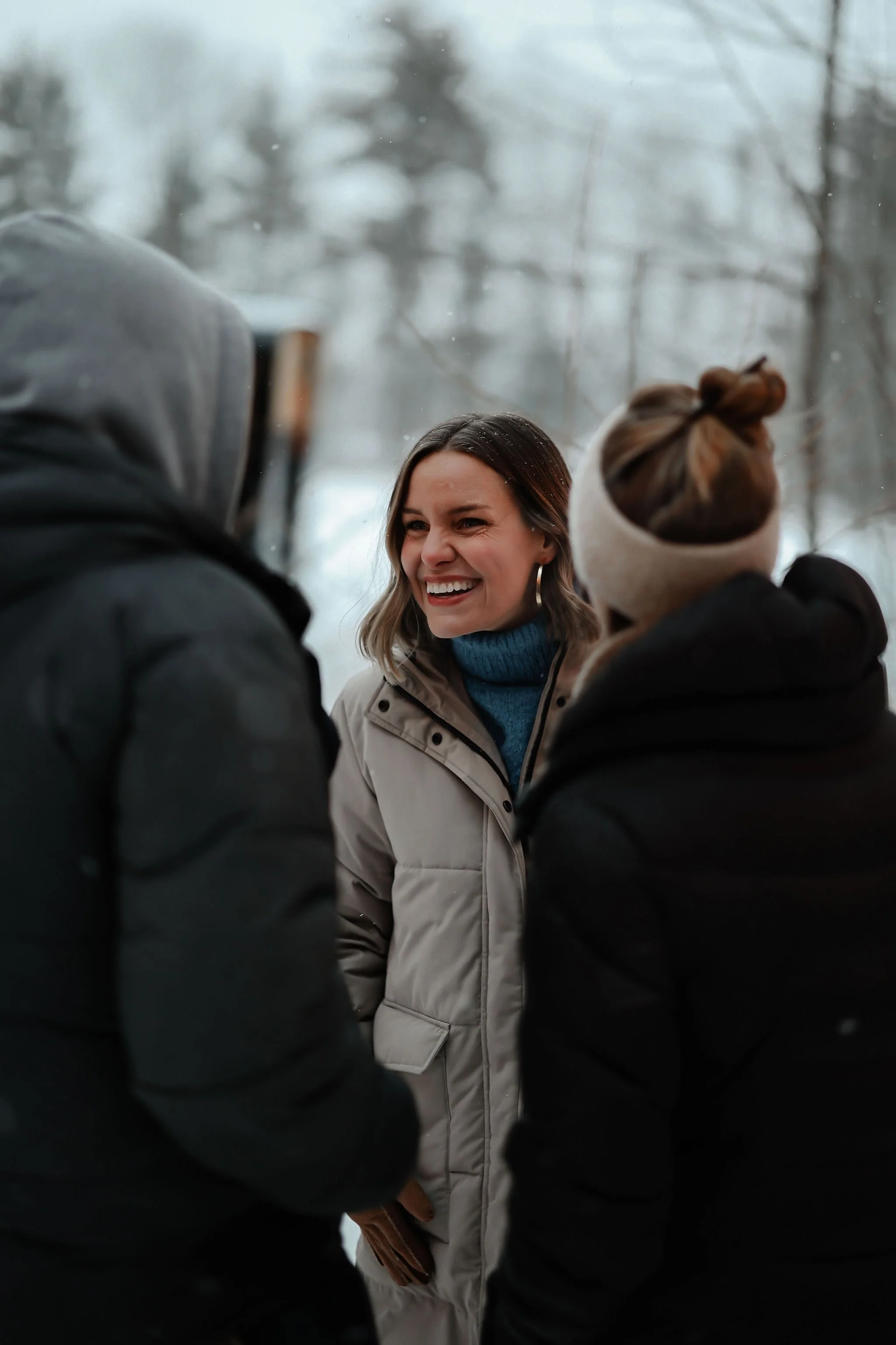 A woman smiling and talking outdoors with two people dressed warmly in winter, trees in the background.