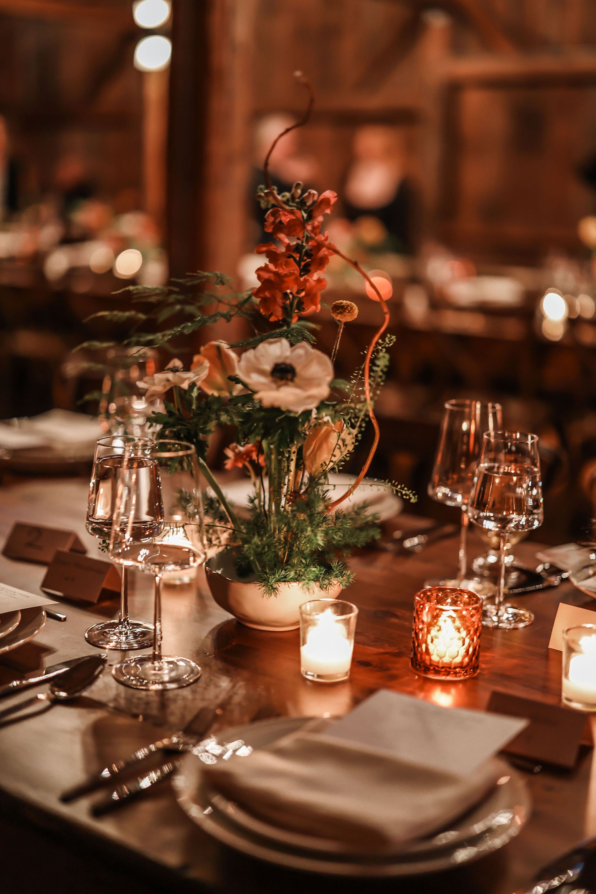 A decorated table with floral centerpiece, candles, wine glasses, and place settings in a dimly lit, rustic setting.
