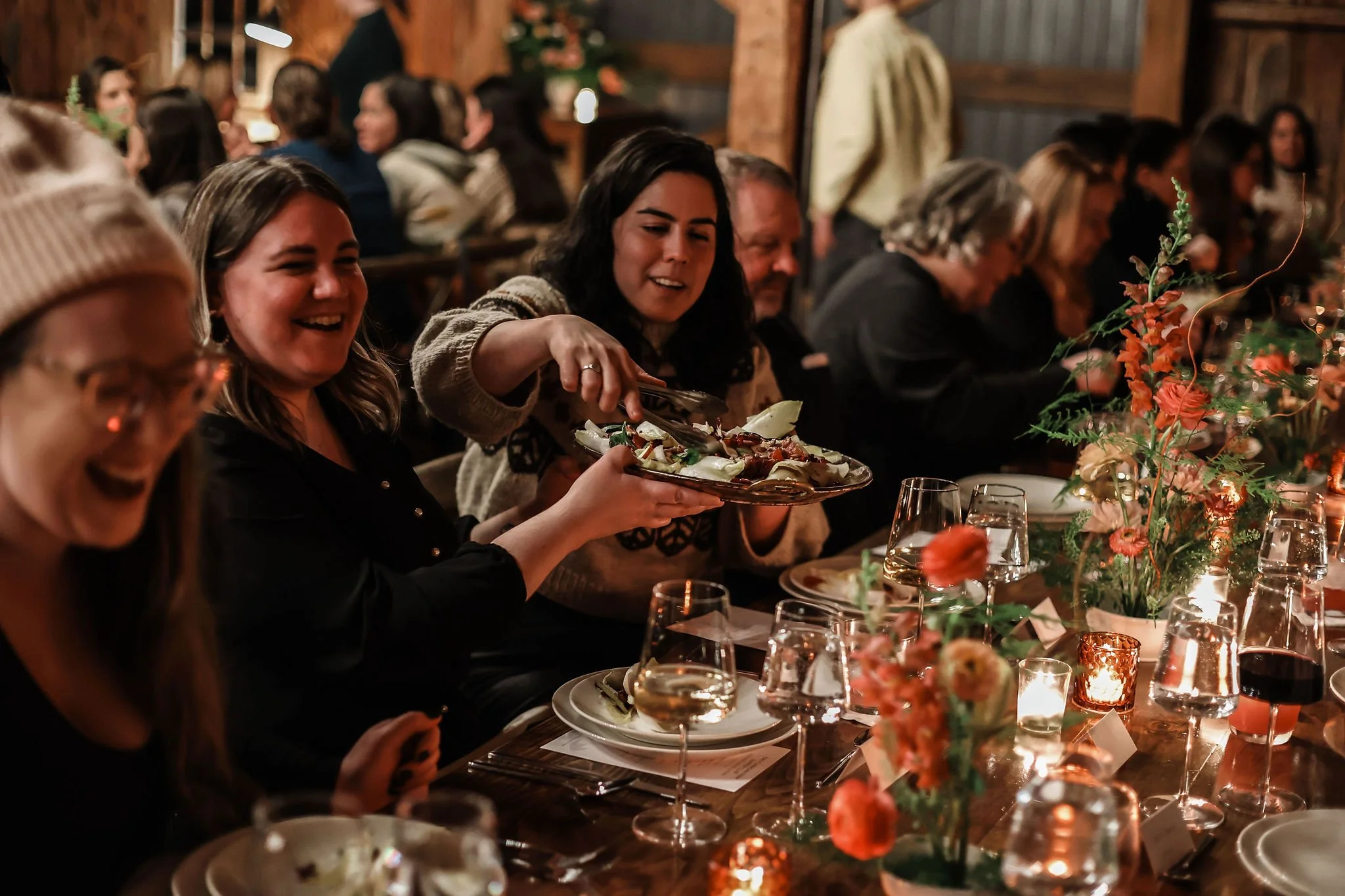 People enjoying a dinner party in a cozy, rustic setting with dim lighting, floral centerpieces, and glasses of wine on the table. One woman is serving food while others laugh and converse.