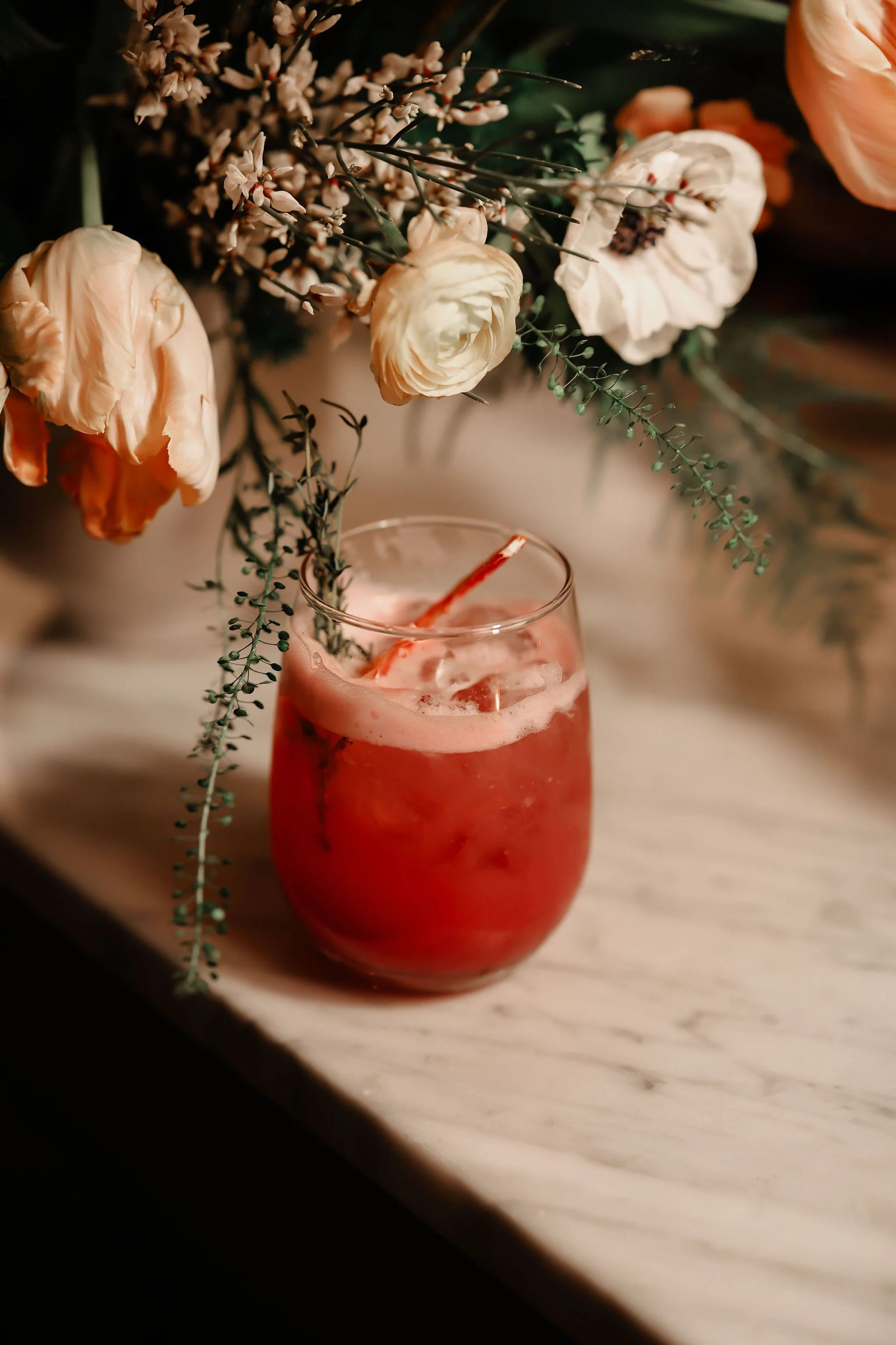 A glass of pink cocktail with ice, garnished with sprigs of flowers, placed on a marble surface under a large bouquet of pale peach and white flowers with greenery.