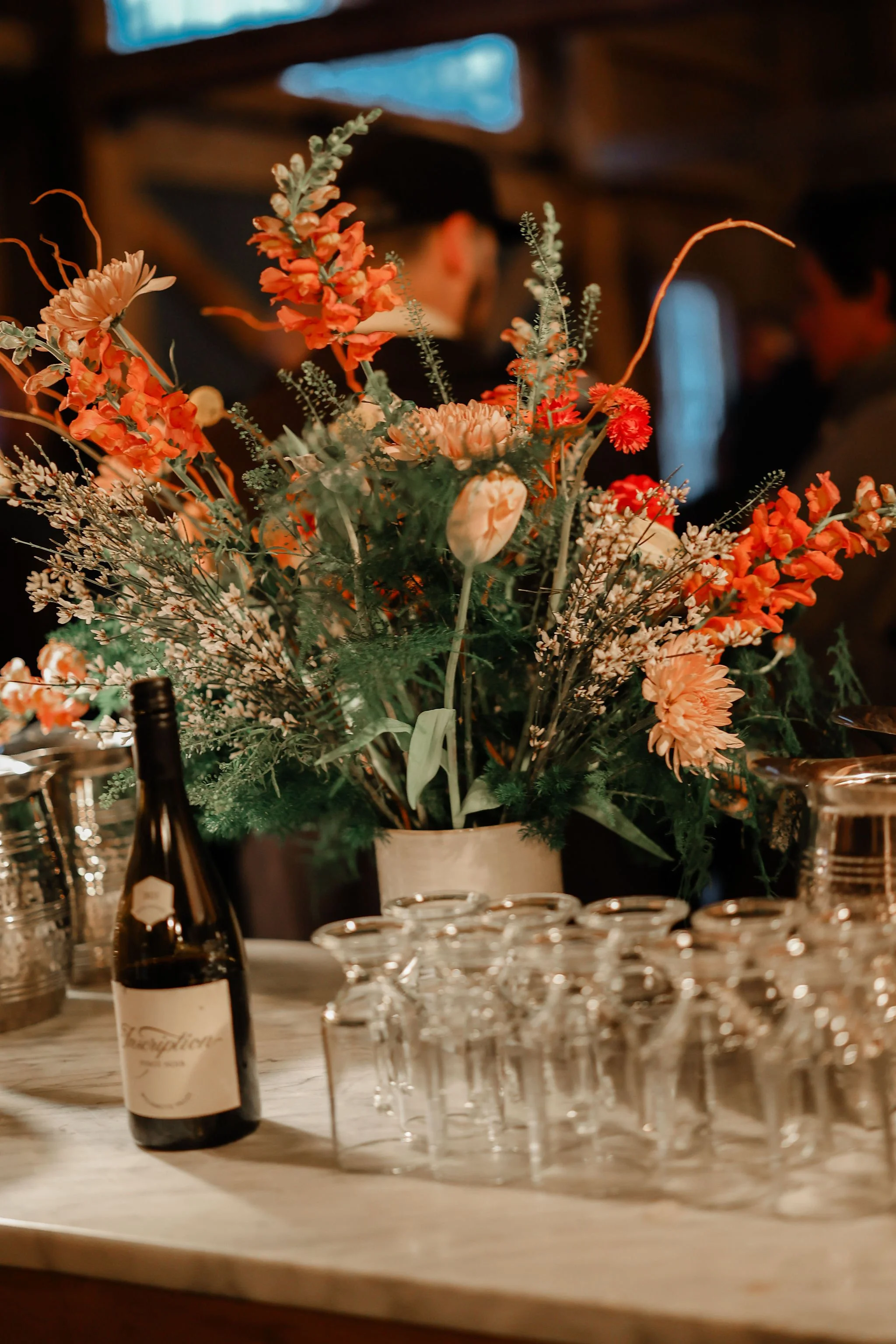 A floral centerpiece with pink, white, and red flowers on a table, surrounded by upside-down wine glasses and a bottle of wine.