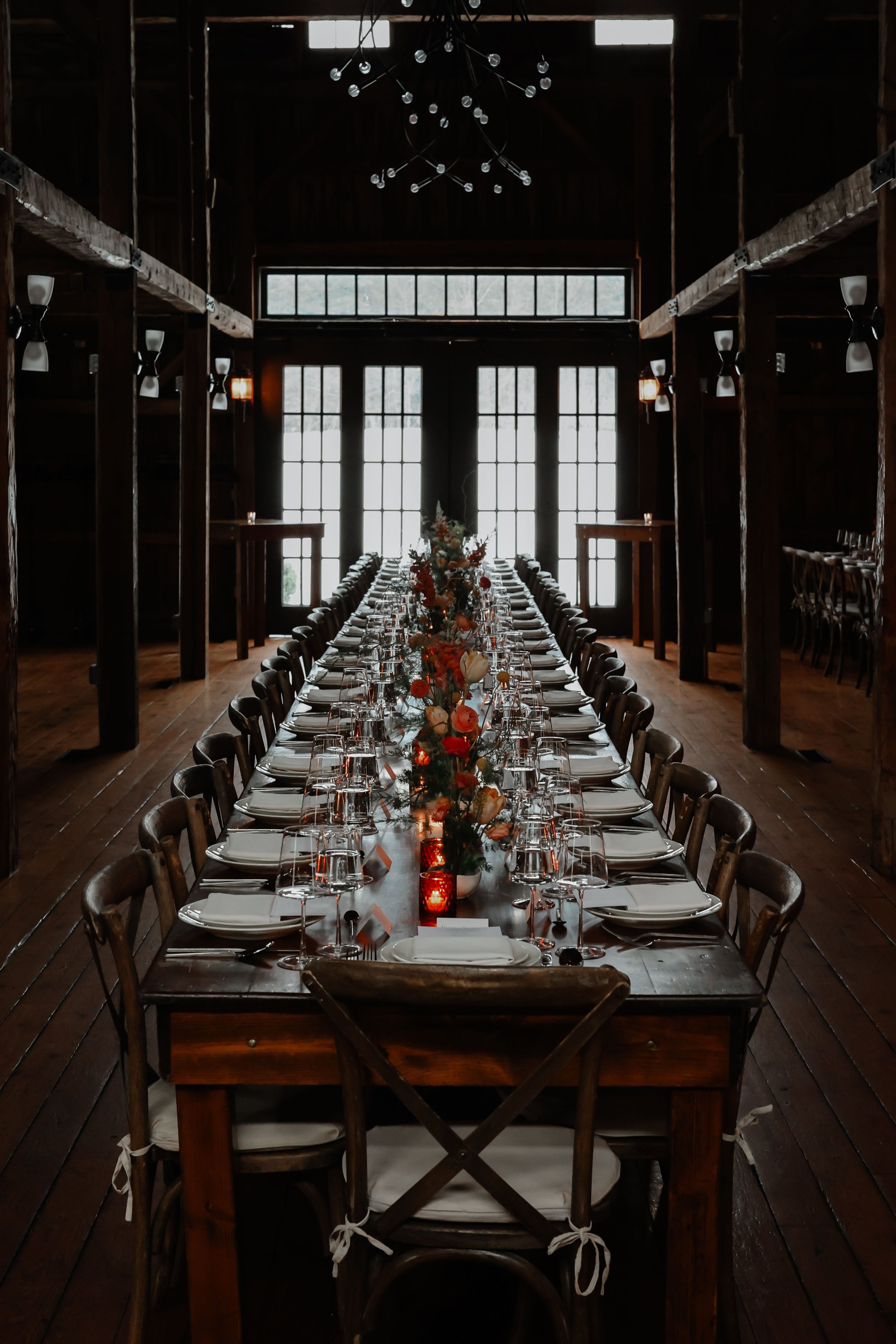 Long dining table set for a formal event inside a rustic wooden venue, decorated with floral centerpieces and candles, with natural light coming through large windows.