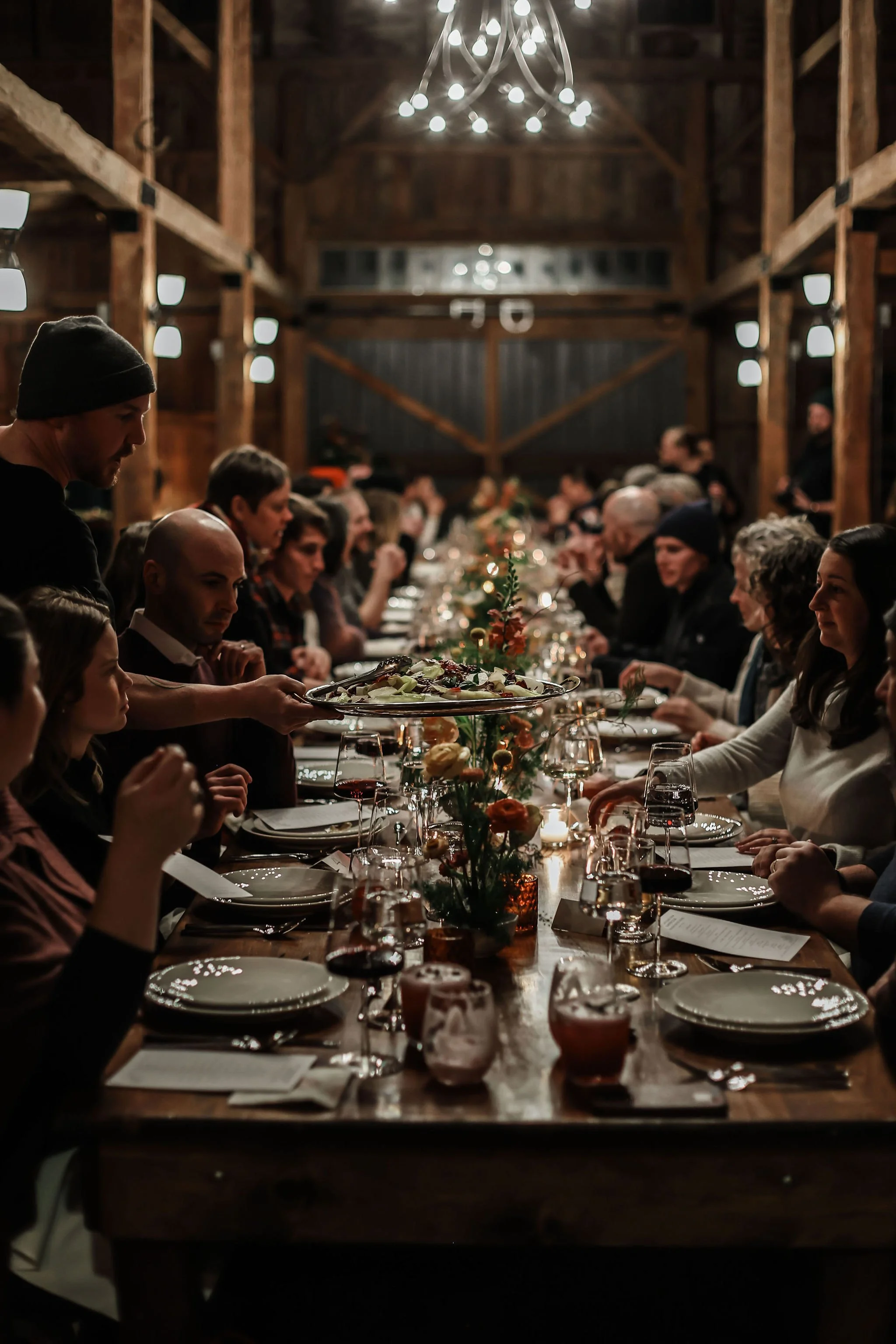 People gathered around a long wooden table in a rustic barn, enjoying a meal during a festive dinner party with warm lighting and decorative centerpieces.