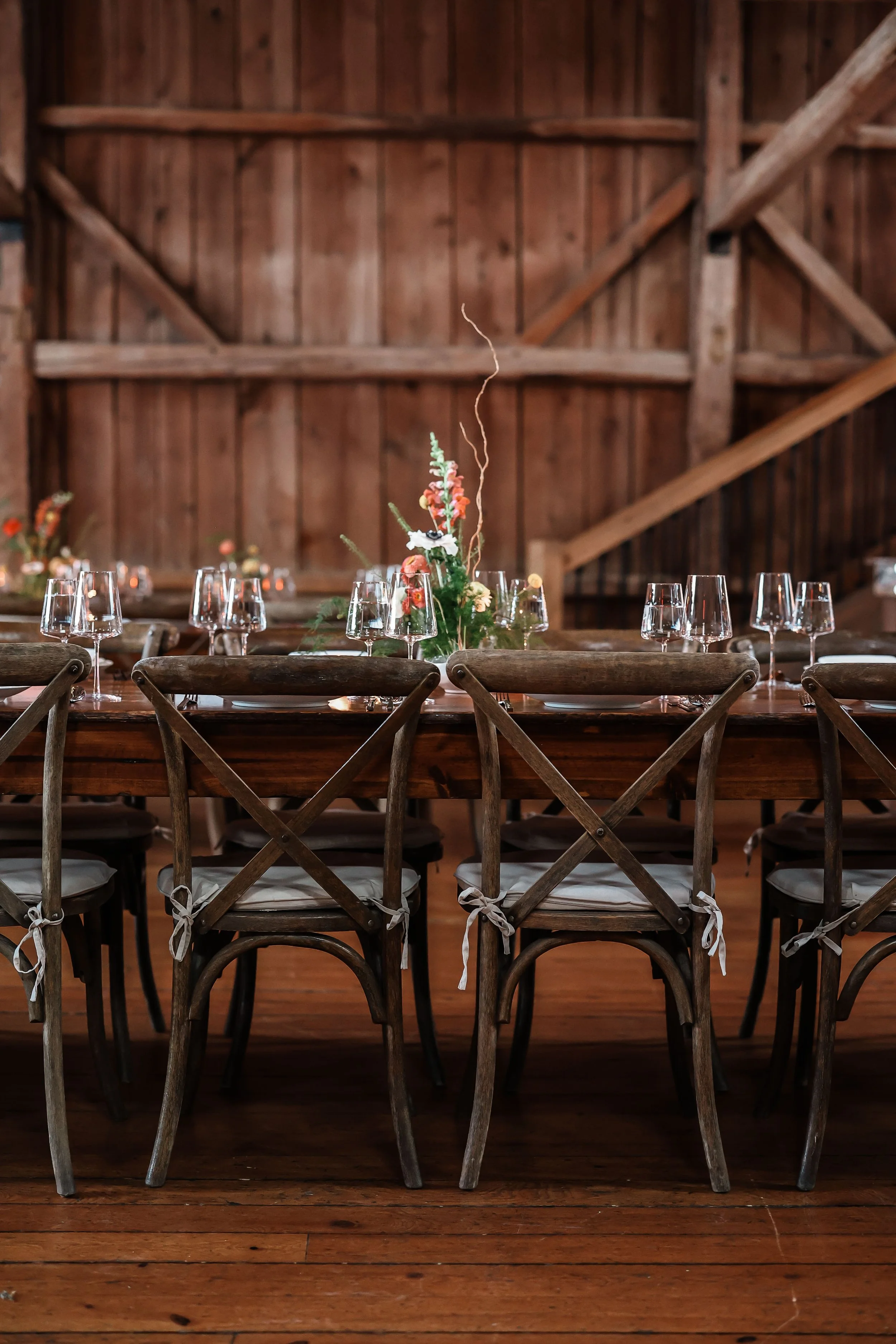 Wooden table set for a meal with glassware and a floral centerpiece inside a rustic barn with wooden walls and ceiling beams.