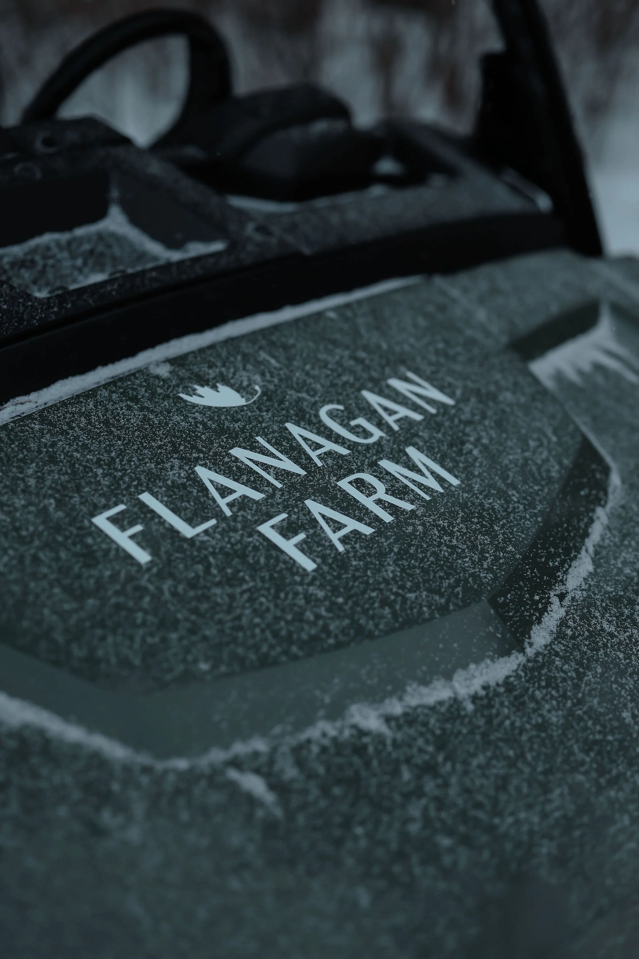 Close-up of a snow-covered sign on a vehicle that reads 'Flanagan Farm' with a mountain logo above the text.