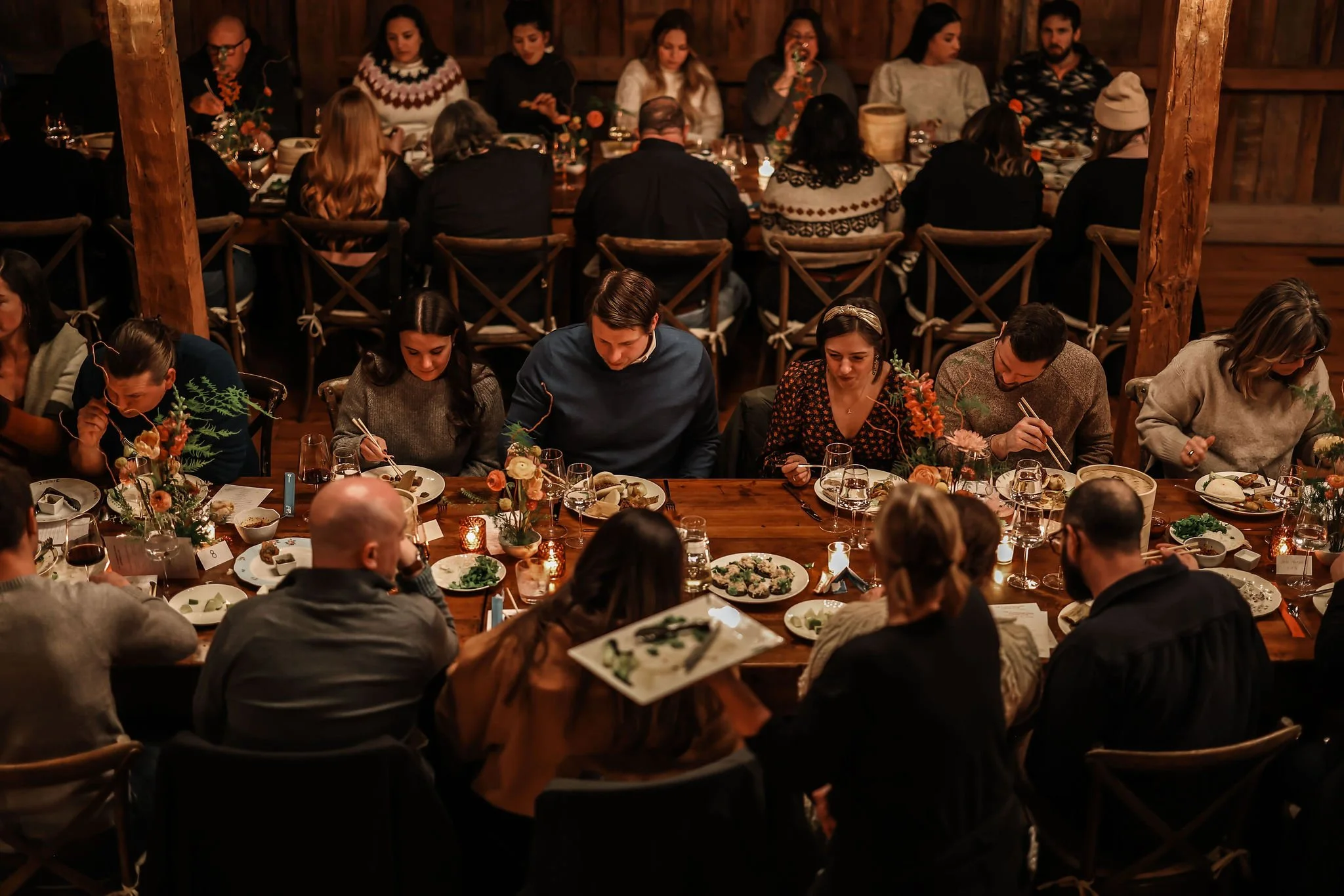 People dining together at a rustic restaurant, sharing a meal with various dishes and floral centerpieces on the table, in a cozy, warmly lit setting.