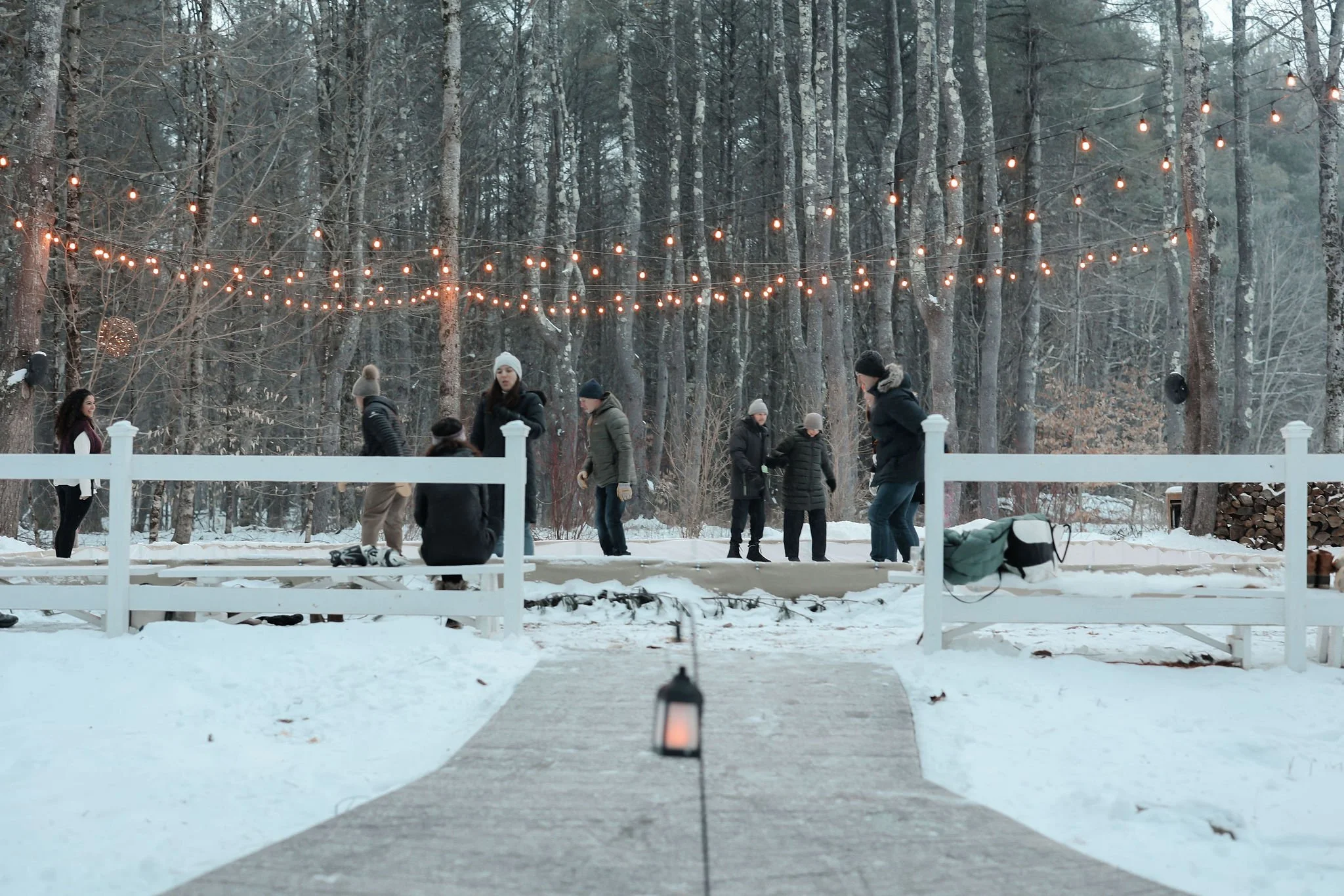 People gathered outdoors in a snowy wooded area decorated with string lights hanging between trees, with a bench and a lantern on the pathway.