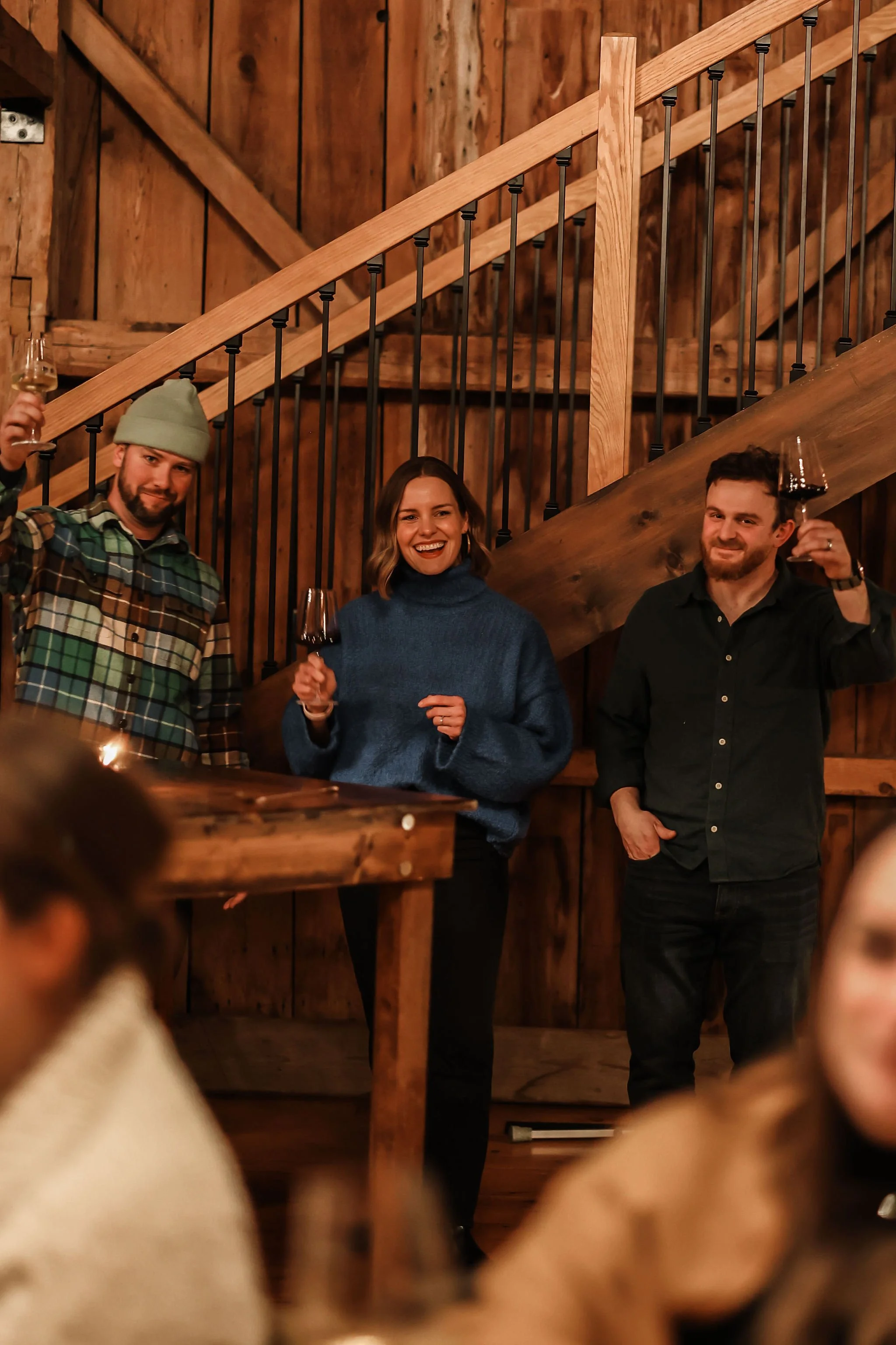 Three people standing inside a rustic wooden room, holding glasses of red wine and smiling at the camera during a social gathering.