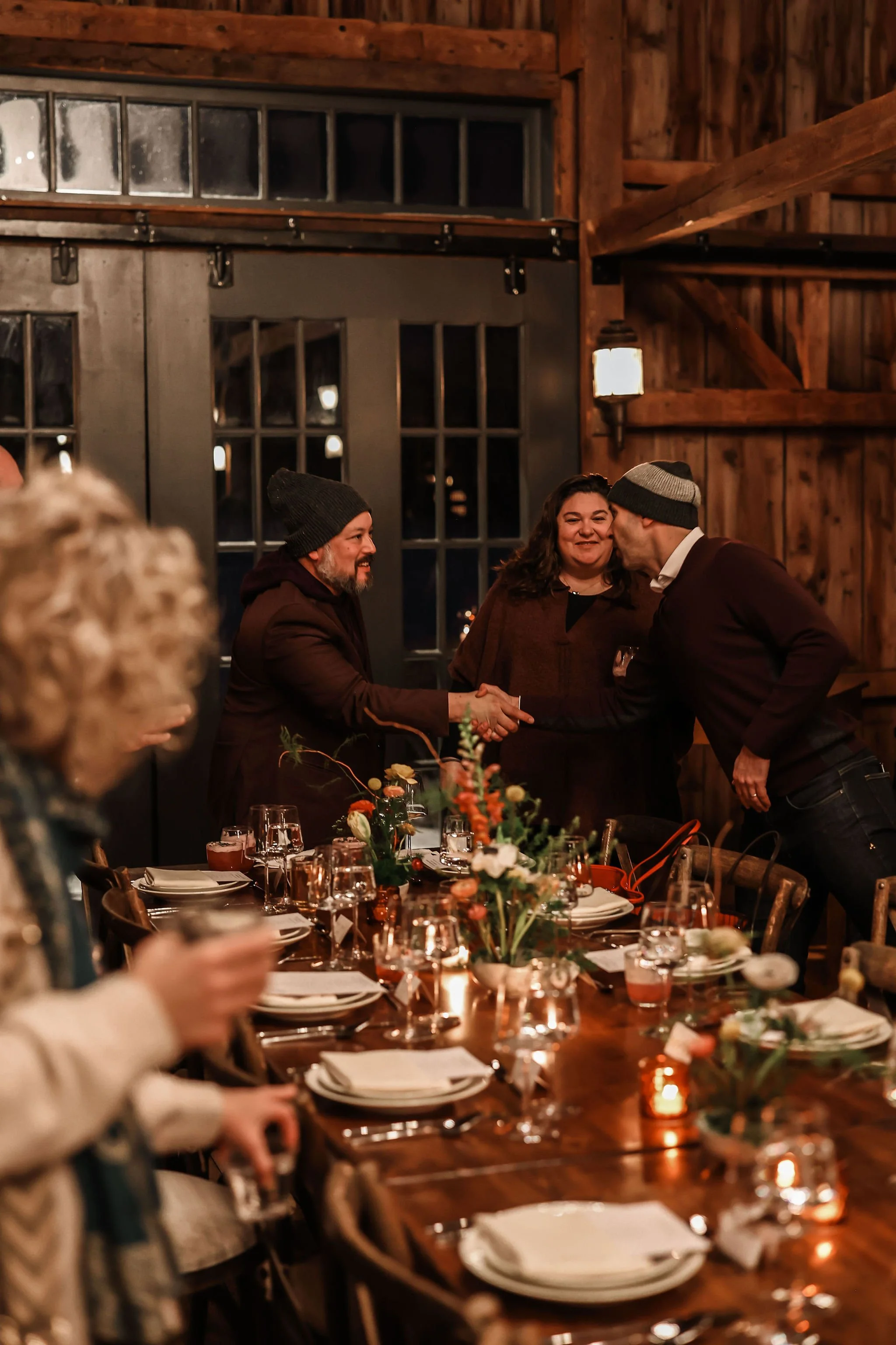 People at a dinner table greeting each other with handshakes and kisses at a warm, rustic wooden indoor celebration.