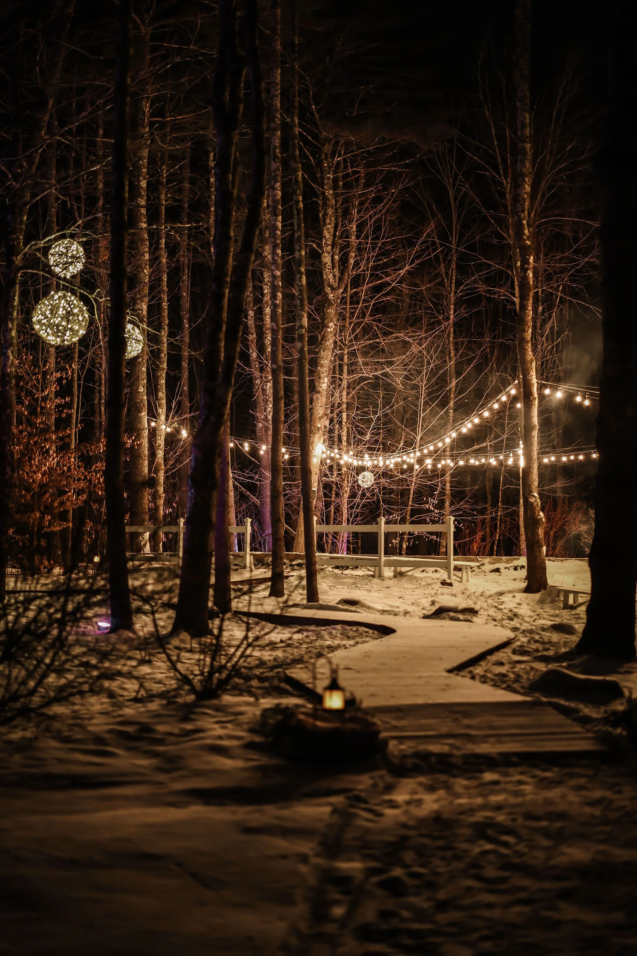 A snowy pathway through a wooded area decorated with hanging string lights and spherical light ornaments, creating a cozy outdoor scene at night.