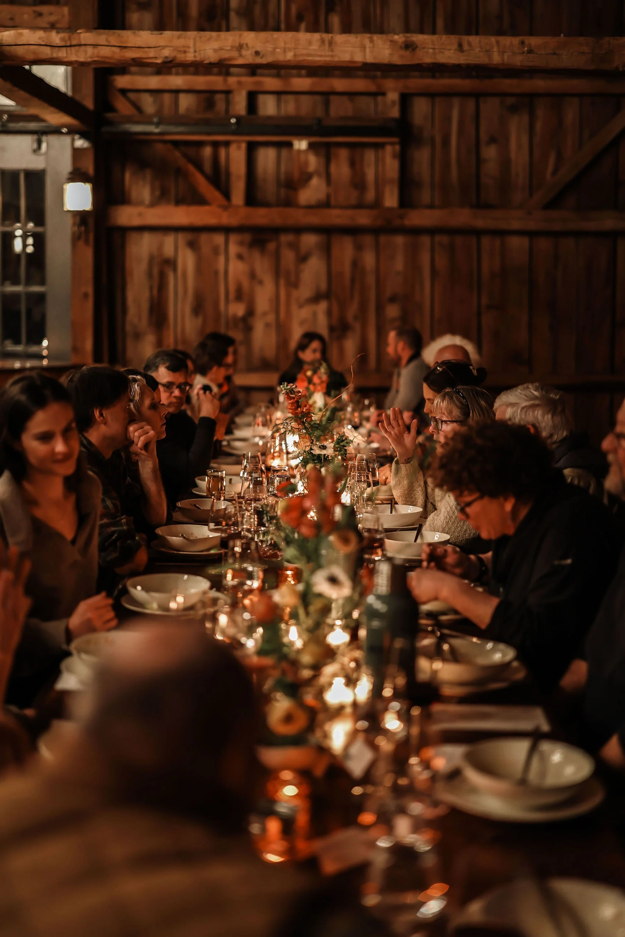 A long dinner table with people dining, decorated with flowers and lit by candles in a rustic wooden barn.