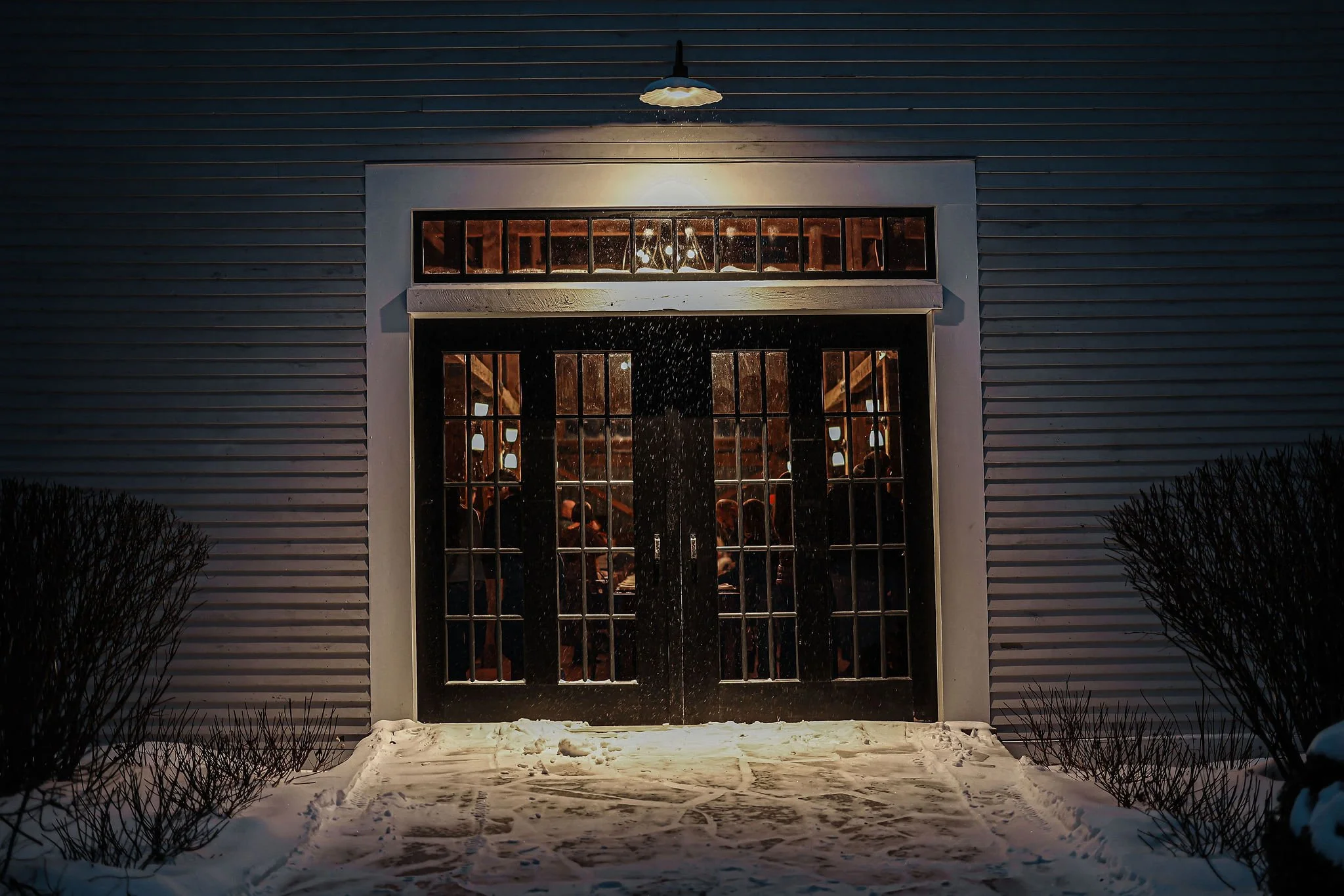 Nighttime view of a barn with large glass double doors and a window above, illuminated by exterior lights. Through the doors, people are visible inside, possibly at a social gathering. Snow covers the ground outside.