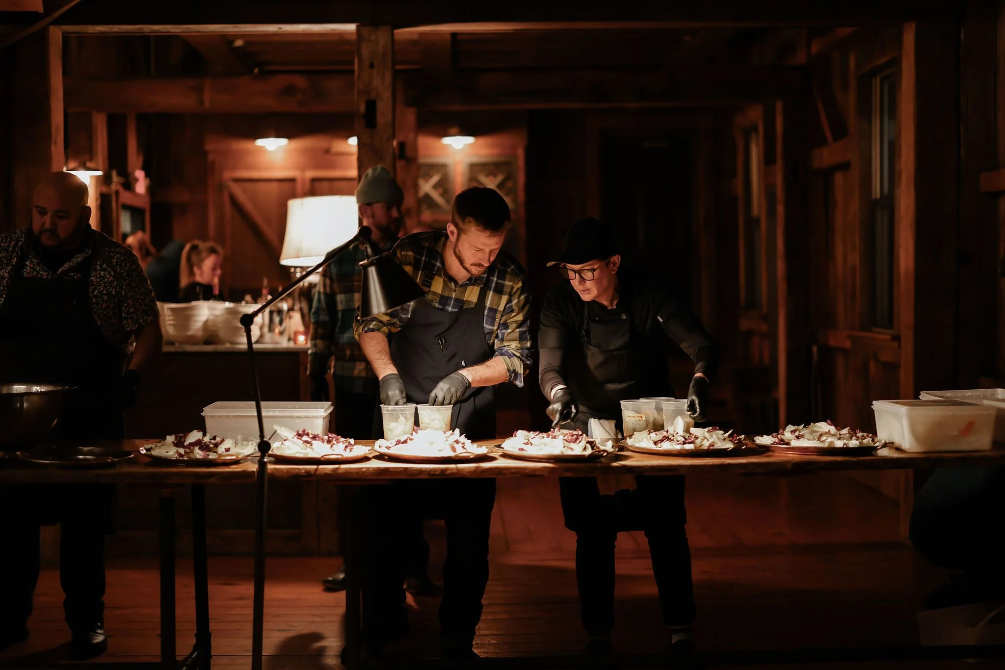 Two chefs preparing food at a wooden table in a rustic, dimly lit kitchen or dining area, with other people working in the background.