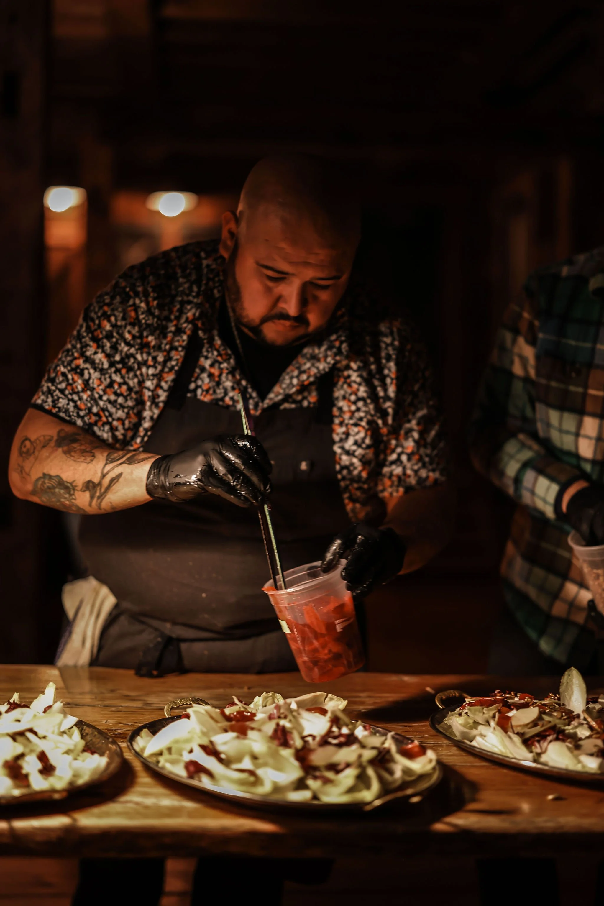 A man with tattoos wearing a floral shirt, black gloves, and an apron preparing food in a dimly lit setting. He is filling plates of what appears to be sliced cabbage or salad with a red sauce. Another person in a plaid shirt is partially visible on 