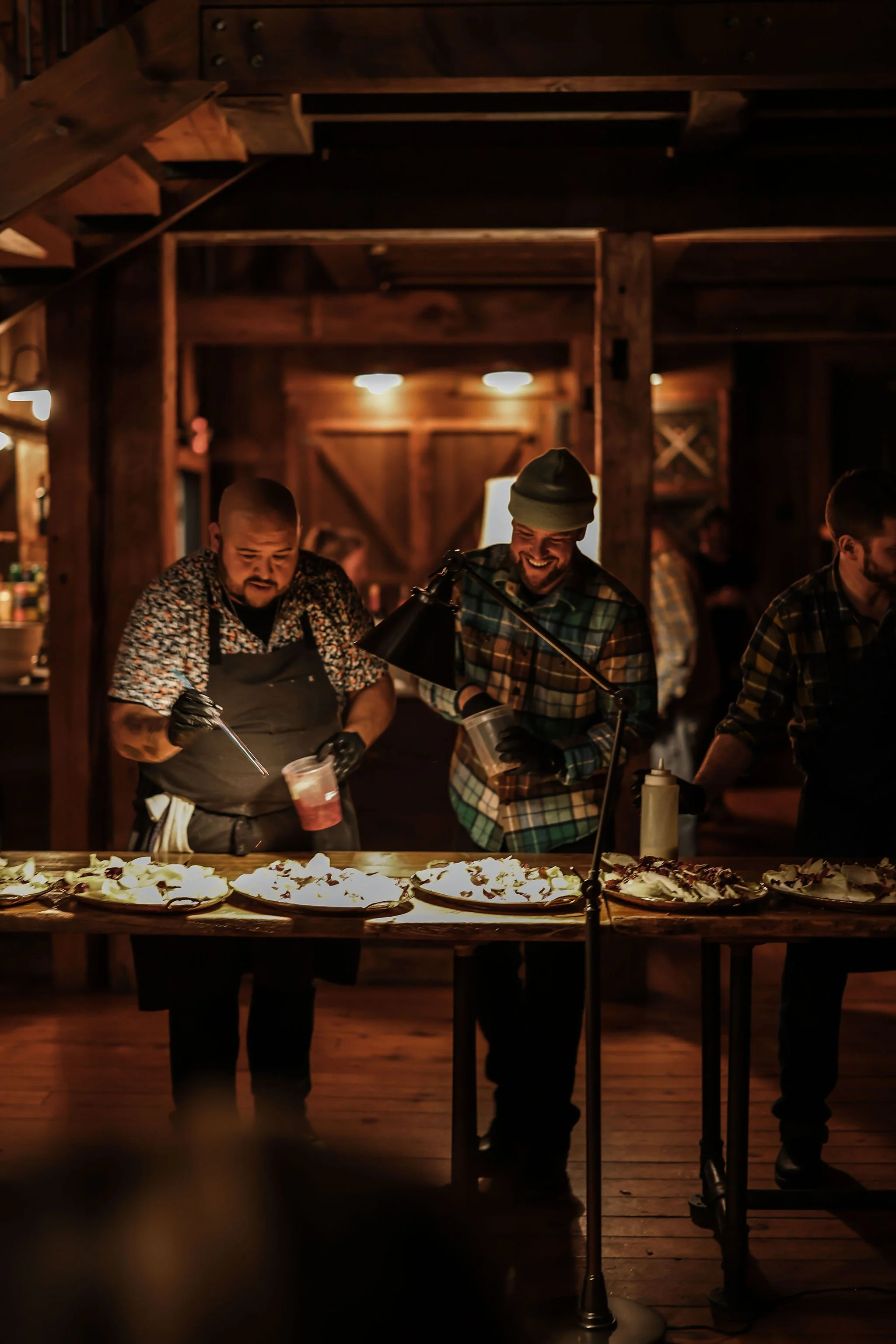 Two men behind a table of food at a rustic indoor event, one wearing a floral shirt and apron, and the other in a plaid shirt and beanie, both smiling, with warm lighting and wooden interior evident.