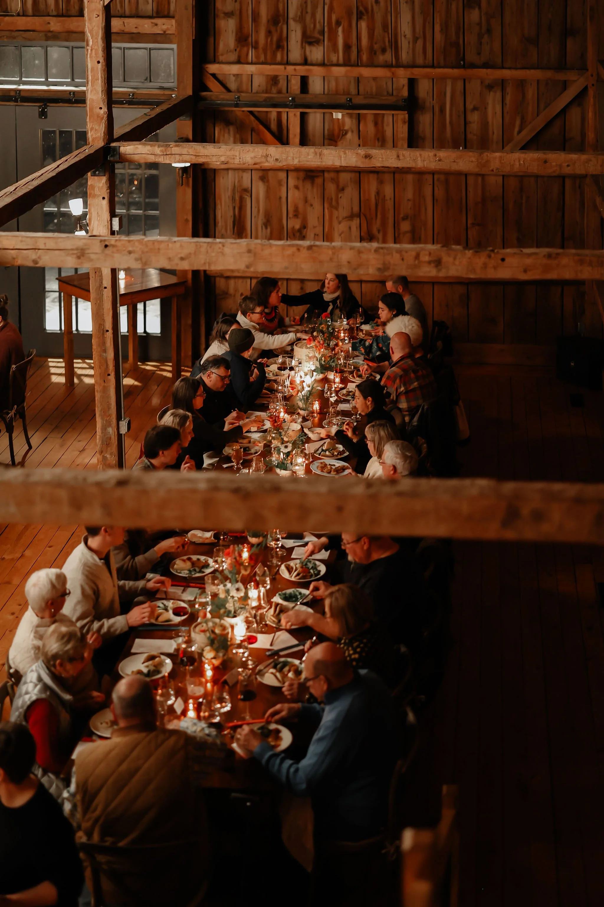 People dining at a long, festively decorated dinner table in a rustic wooden barn with warm lighting.