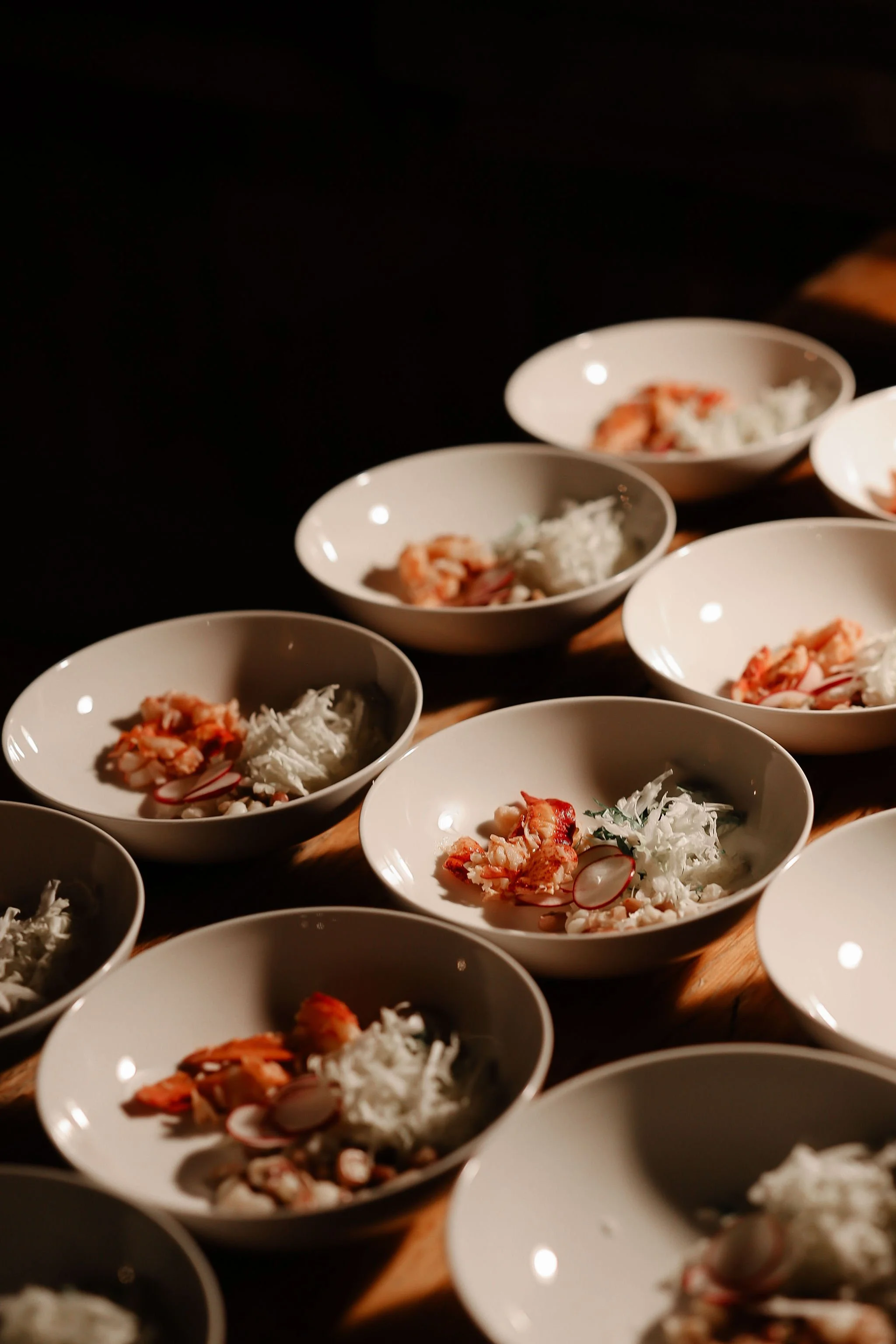 Multiple white bowls filled with seafood, radish slices, and shredded white vegetables arranged on a wooden tray under low lighting.