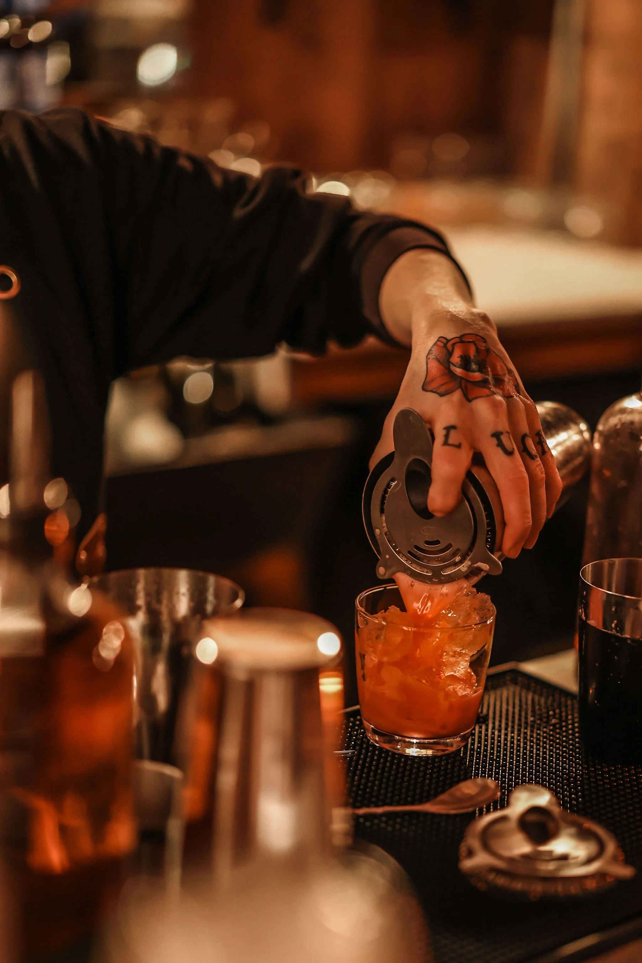 Bartender with tattoos pouring a drink over ice in a glass behind a bar at night.