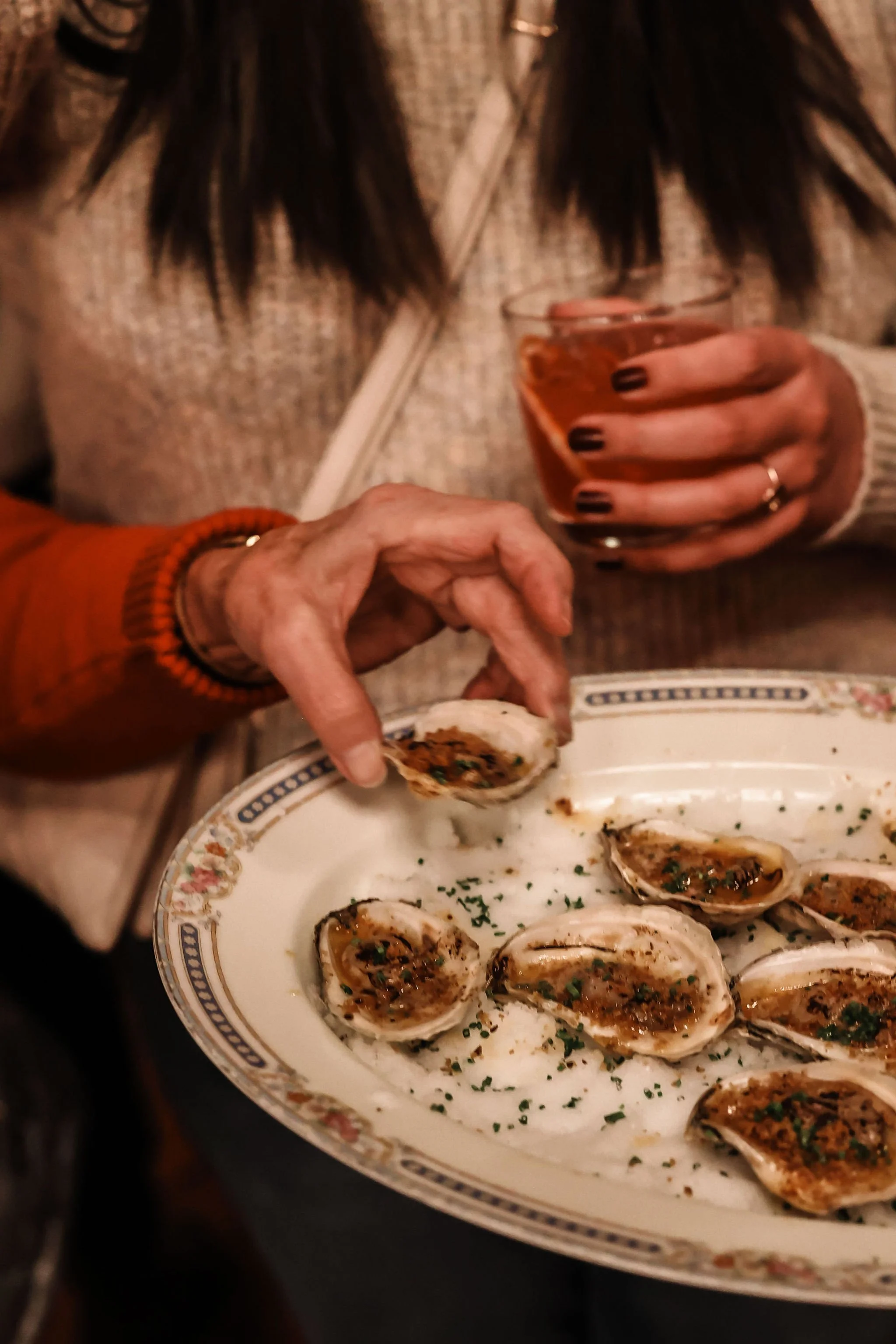 A person with dark hair, wearing a beige sweater and a red bracelet, holding a glass of rosé wine in one hand and a oyster in the other, with a plate of oysters garnished with herbs in front of them.