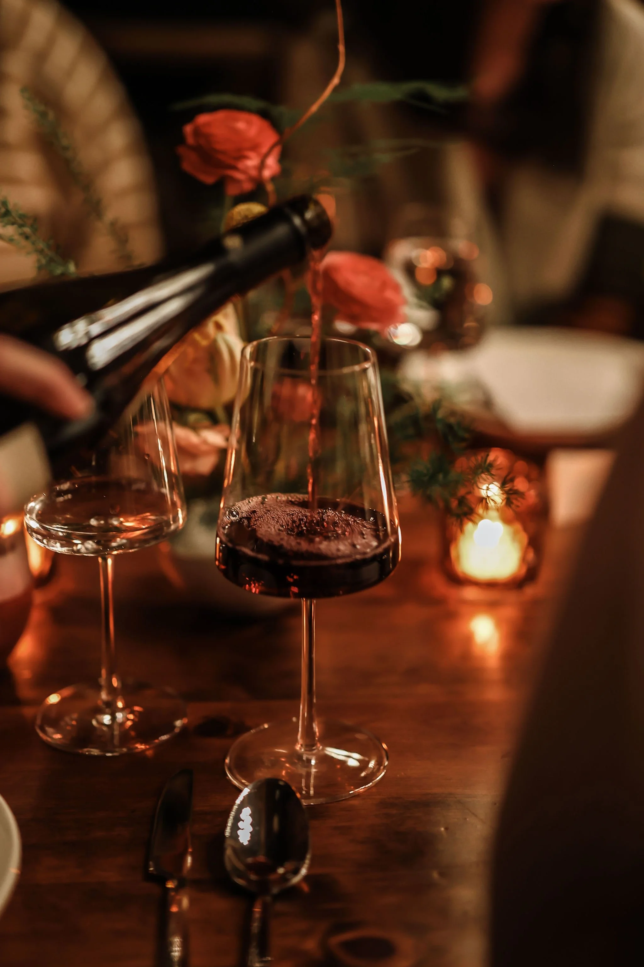 Pouring red wine into a glass at a dimly lit table with flowers and candles.