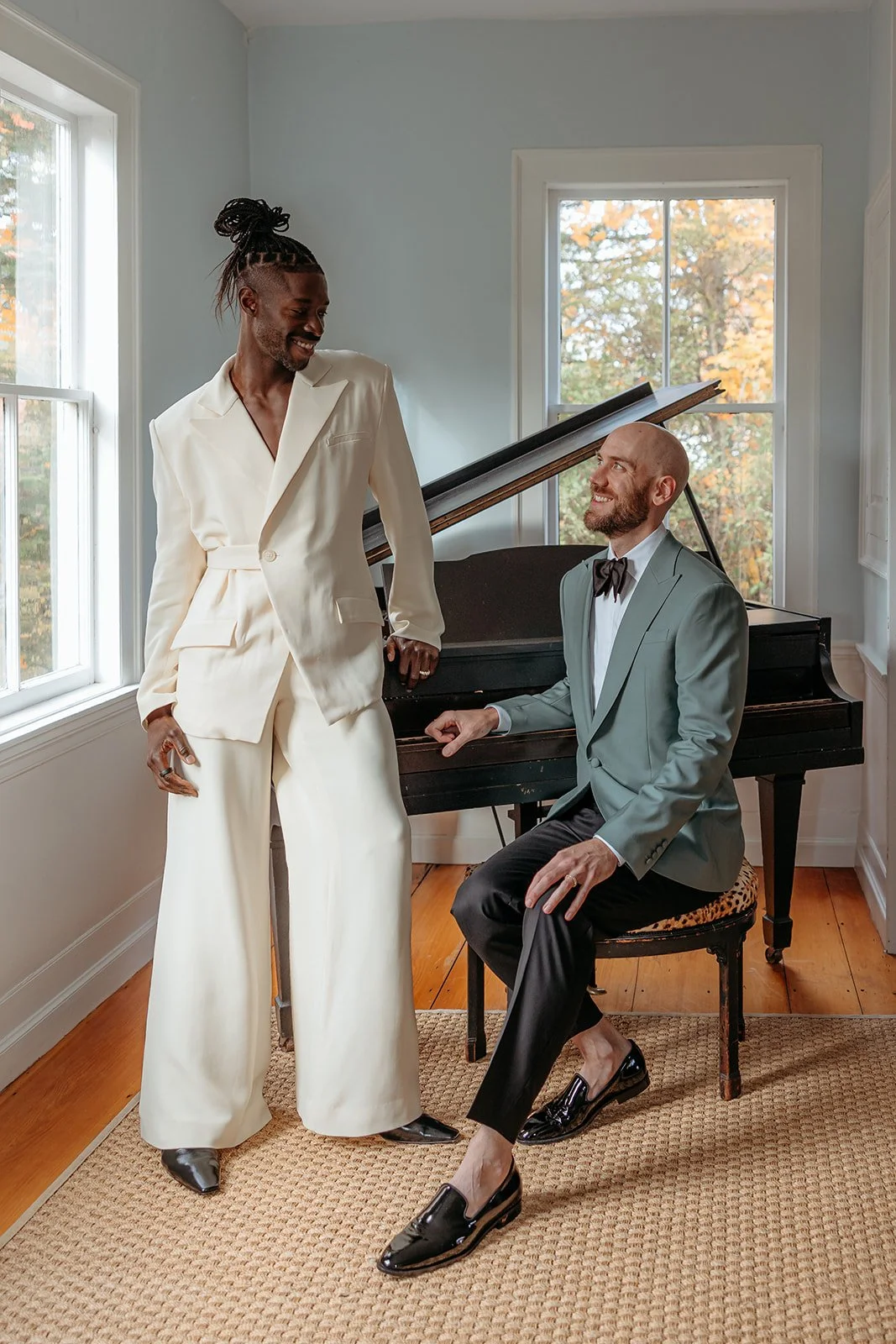 Two men in formal attire, one standing and the other sitting, near a black grand piano in a bright room with large windows and autumn trees outside.