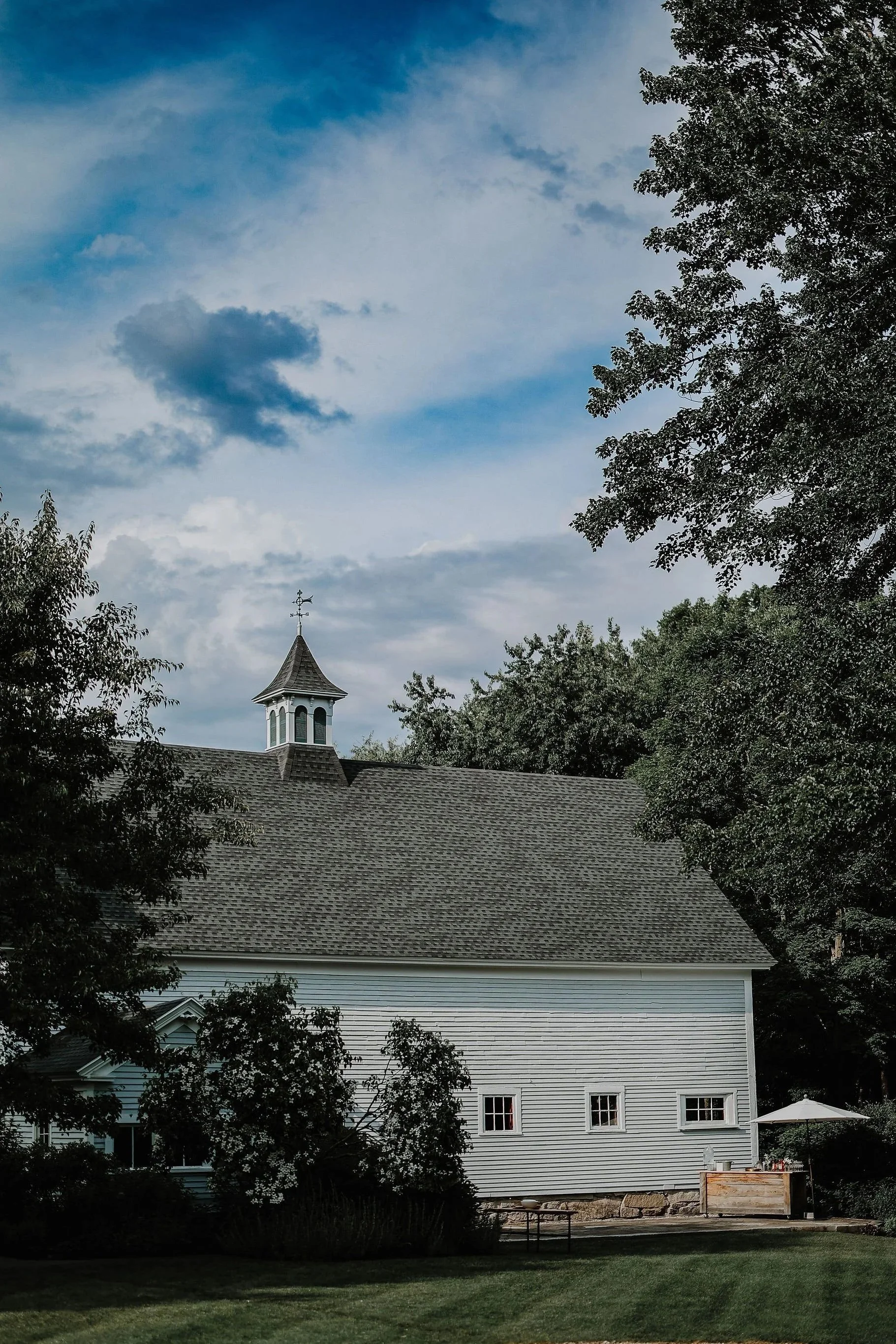 White wooden church with a small steeple, surrounded by trees with a partly cloudy sky overhead.