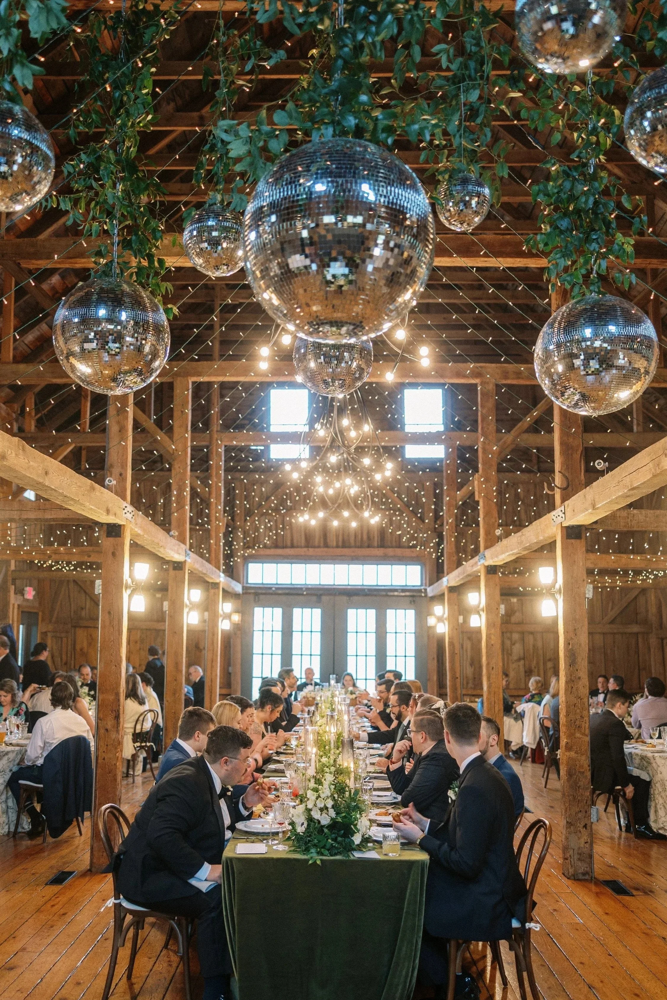 A large gathering of people seated at a long, elegantly decorated dining table inside a rustic wooden barn with string lights and hanging disco balls.
