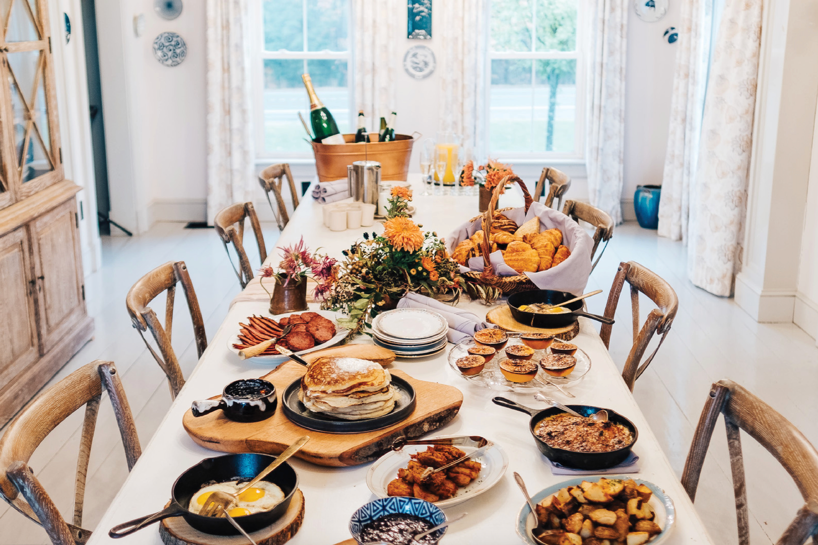 A dining table set with breakfast foods including pancakes, eggs, bacon, muffins, and various dishes, with a floral centerpiece and a basket of croissants. In the background, windows with curtains and a wooden china cabinet are visible.