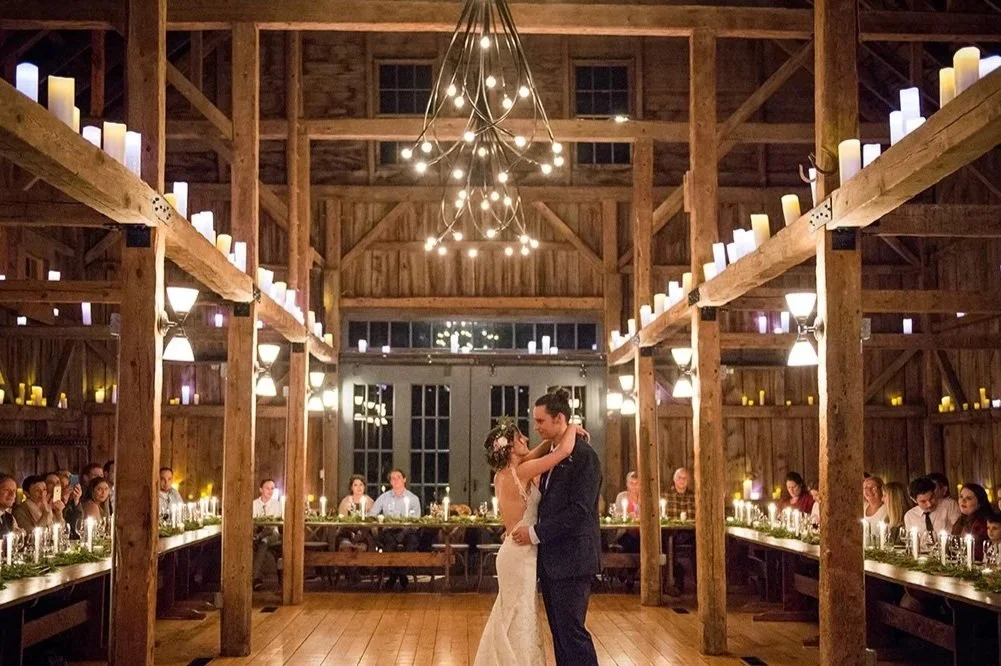 A bride and groom dancing in the center of a rustic wedding reception with wooden walls and ceiling, surrounded by seated guests, candles, and string lights hanging from the ceiling.
