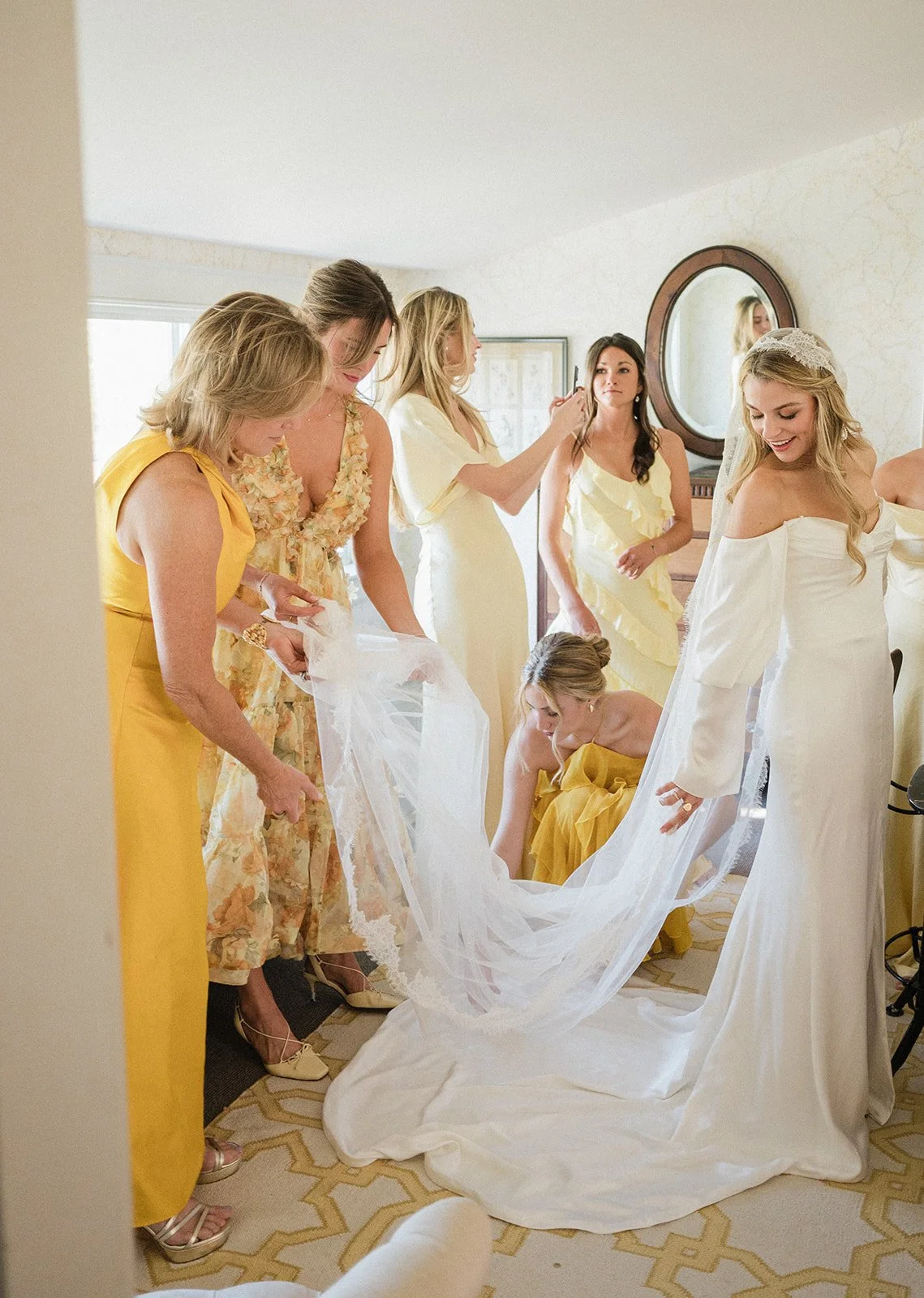 A bride in a white wedding dress is surrounded by her bridesmaids in yellow dresses, helping her with her gown in a bedroom.