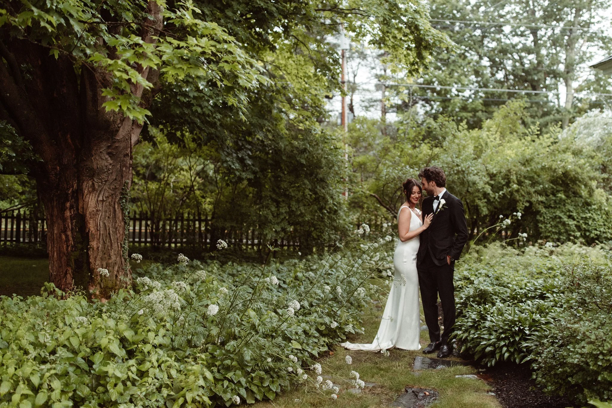 A bride and groom standing together outdoors in a lush garden, smiling and looking at each other, surrounded by green plants and trees, with a large tree on the left and a wooden fence in the background.