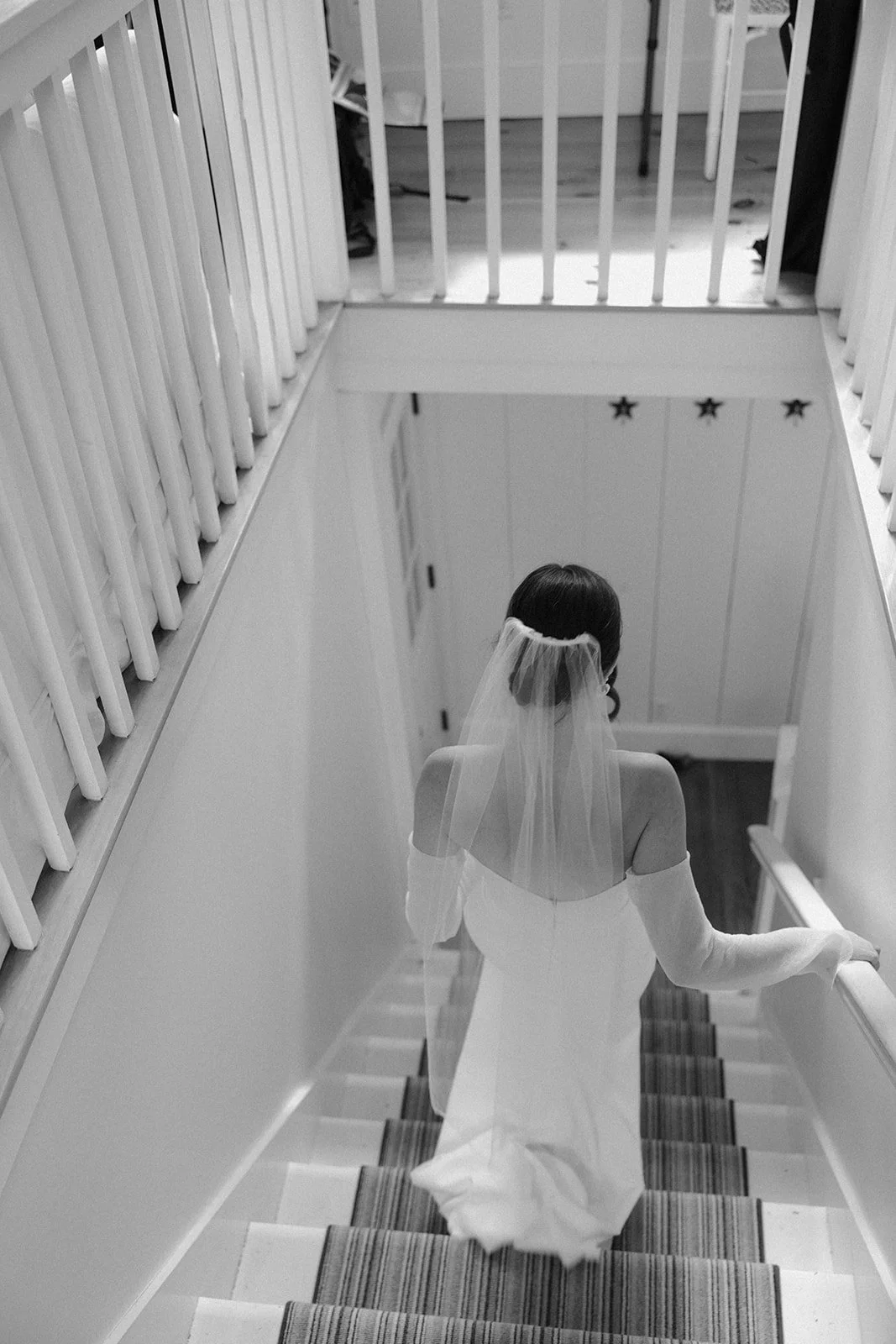 A woman dressed as a bride in a white wedding gown with a veil, descending a staircase with striped carpet, hand on the railing, in a home interior with white walls and star decorations.