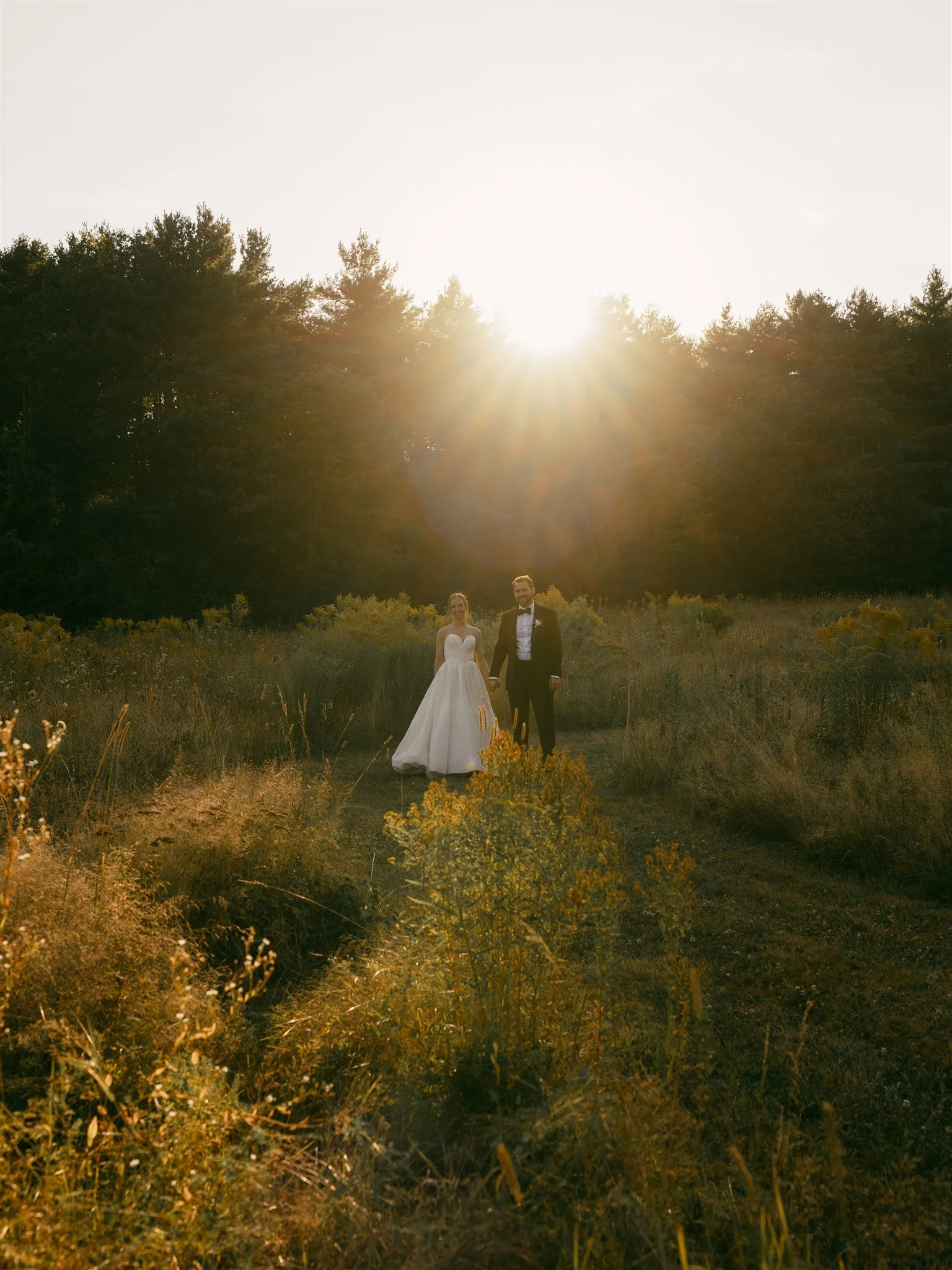Bride and groom holding hands in a field at sunset with trees in the background.