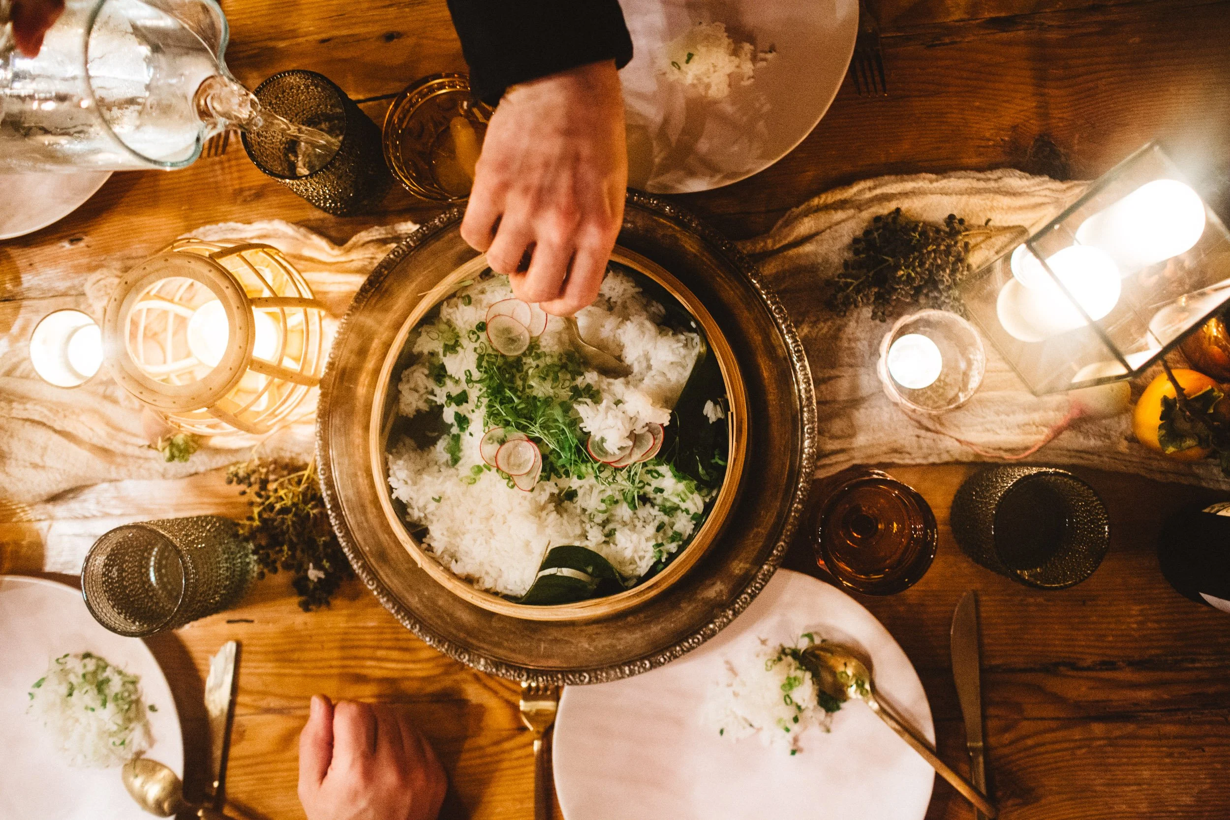 A top-down view of a dinner table with a person serving rice and vegetables from a large bowl, surrounded by plates, glasses, candles, and decorative items.