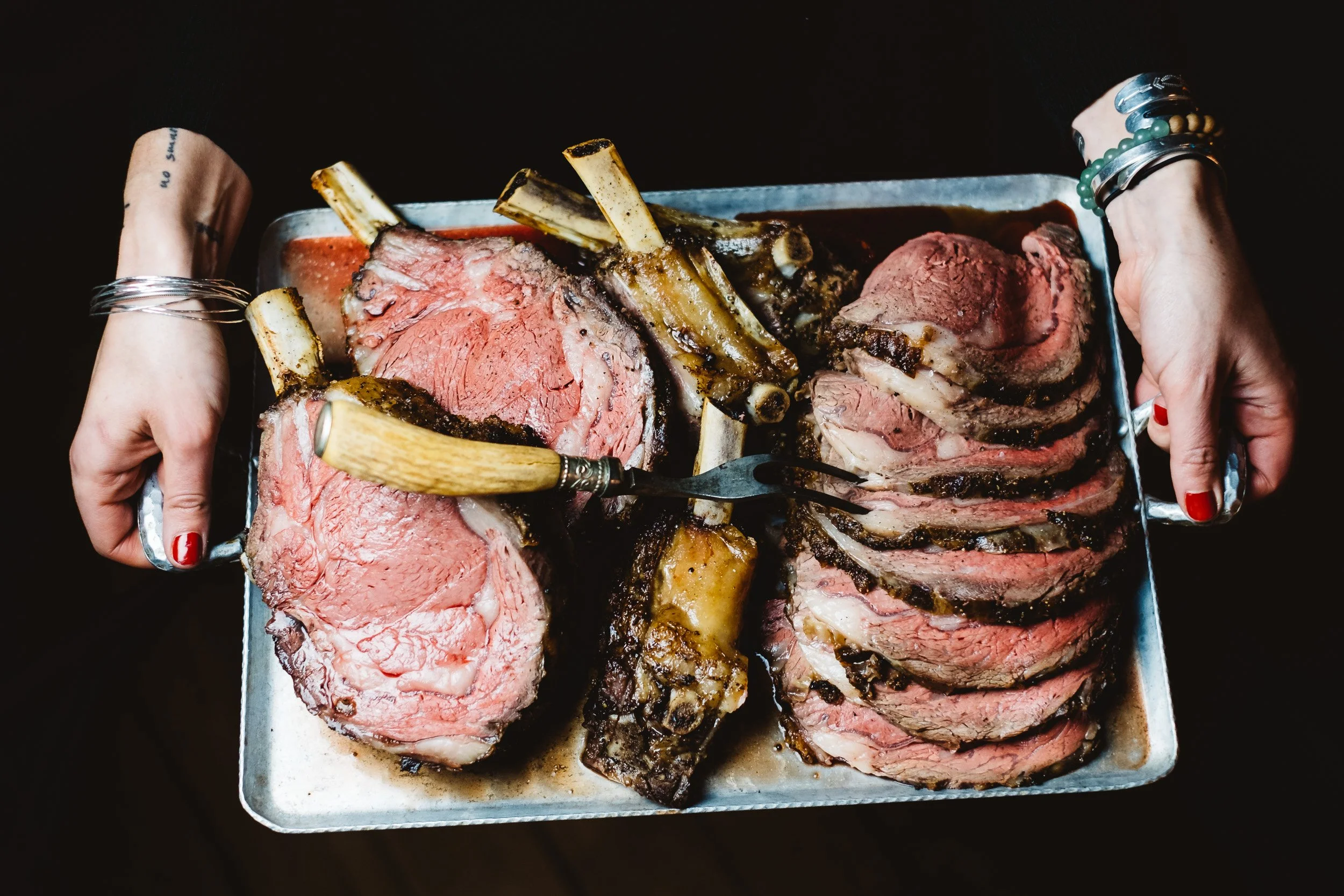 Person holding a tray of sliced medium-rare cooked prime rib with bones, garnished with herbs.