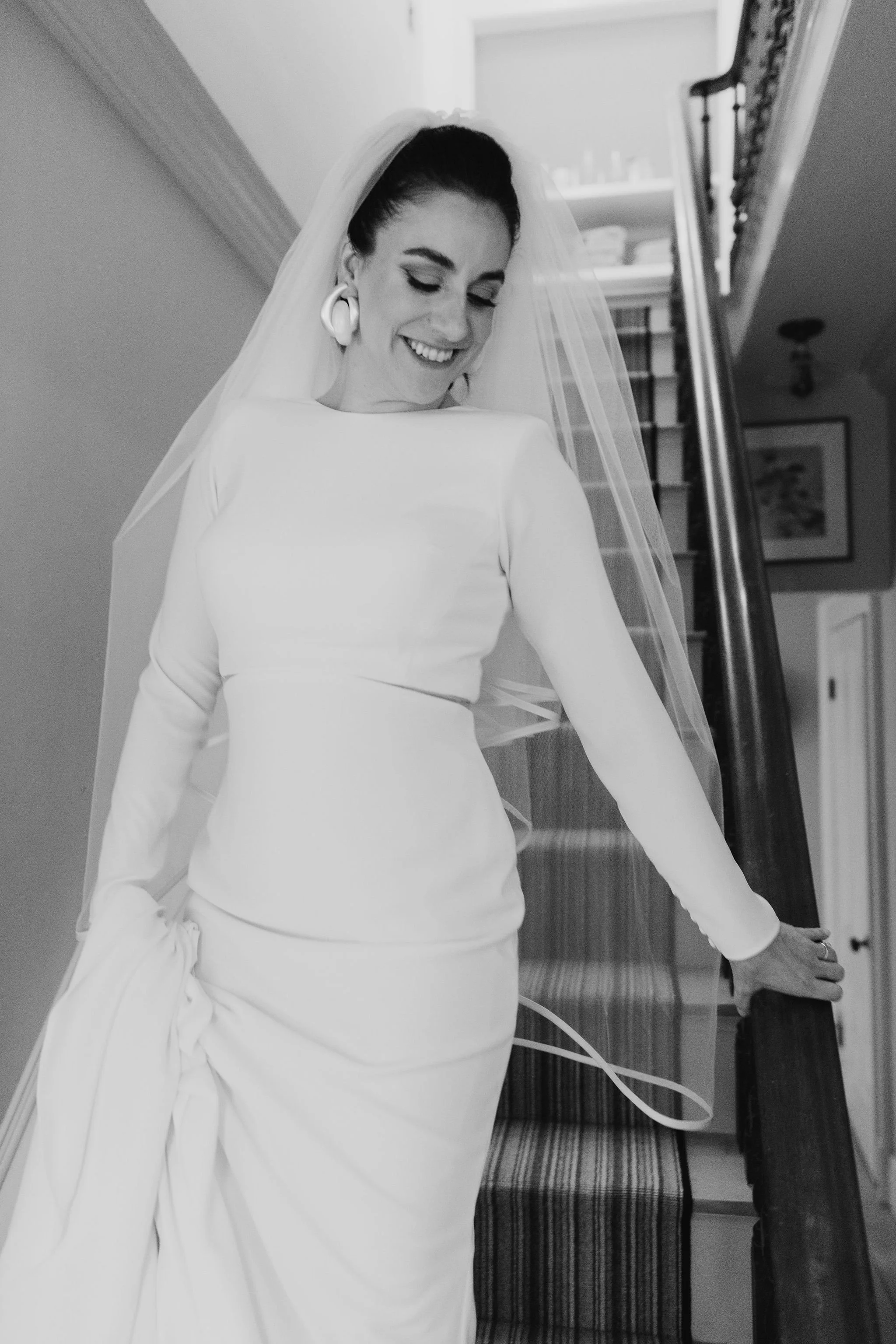 A woman in a wedding dress and veil standing on a staircase, smiling and looking down, holding the stair railing with one hand.