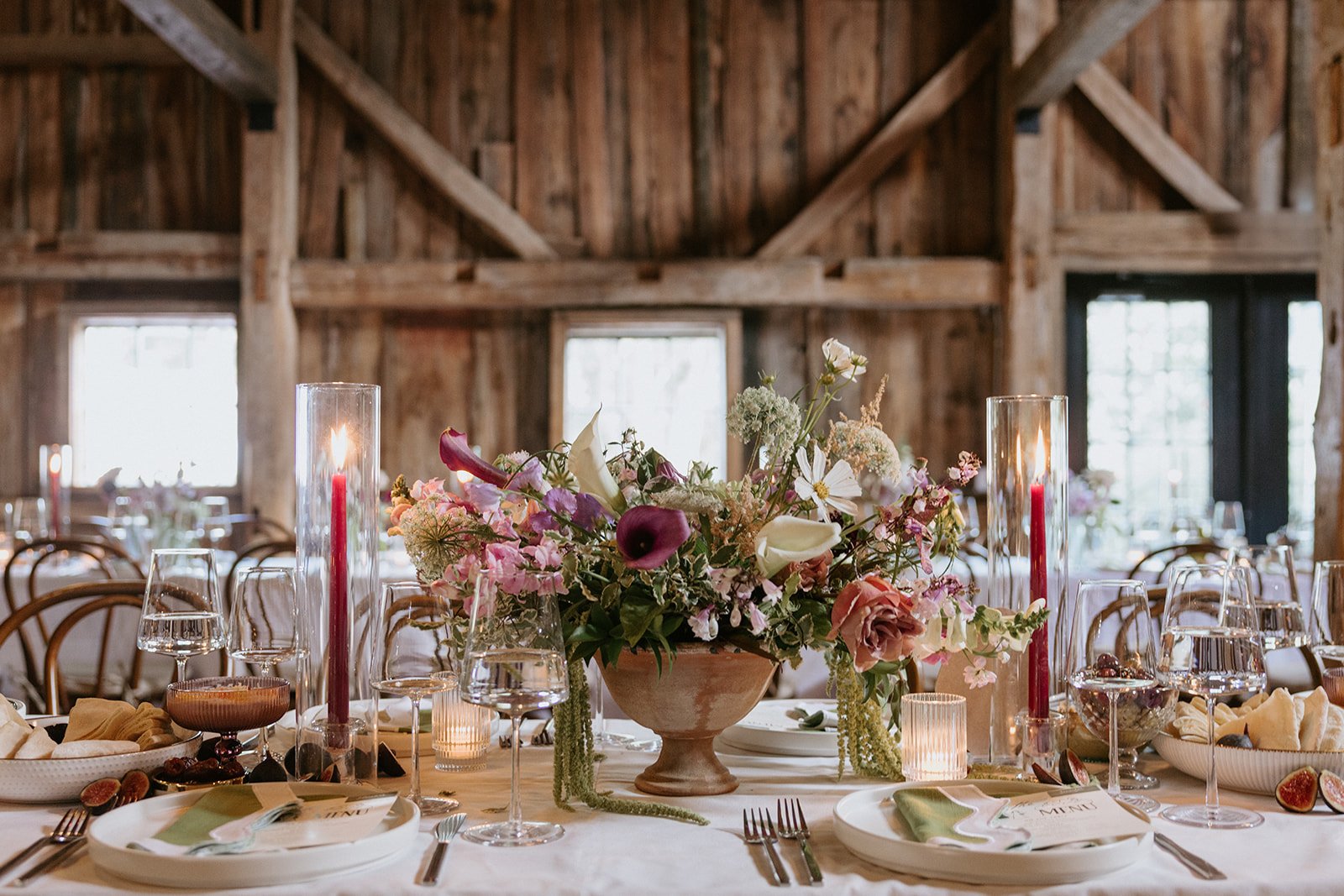 Elegant table setting with a floral centerpiece, candles, wine glasses, and tableware in a rustic wooden barn setting.