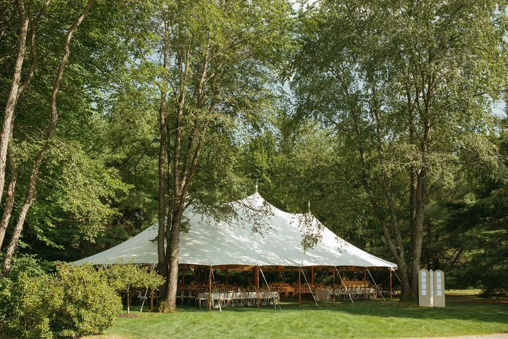 Large white event tent set up outdoors among trees on a grassy area, with tables and chairs inside.