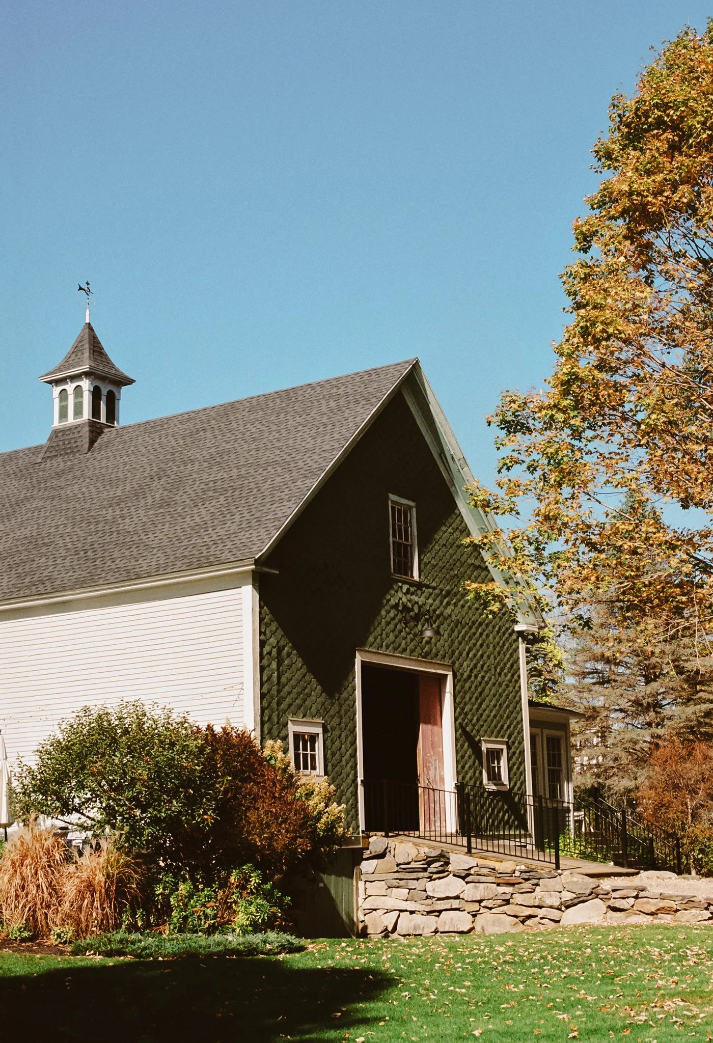 A small church with a green exterior and a brown shingled roof, surrounded by trees with fall foliage, under a clear blue sky.