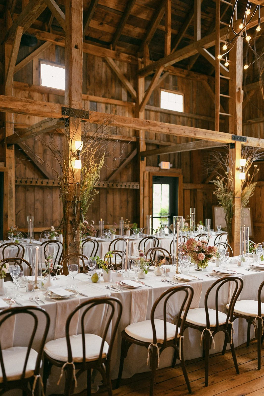 Indoor rustic wedding reception decorated with flowers in vases, tall glass candle holders, and white tablecloths, set inside a wooden barn with exposed beams and windows.