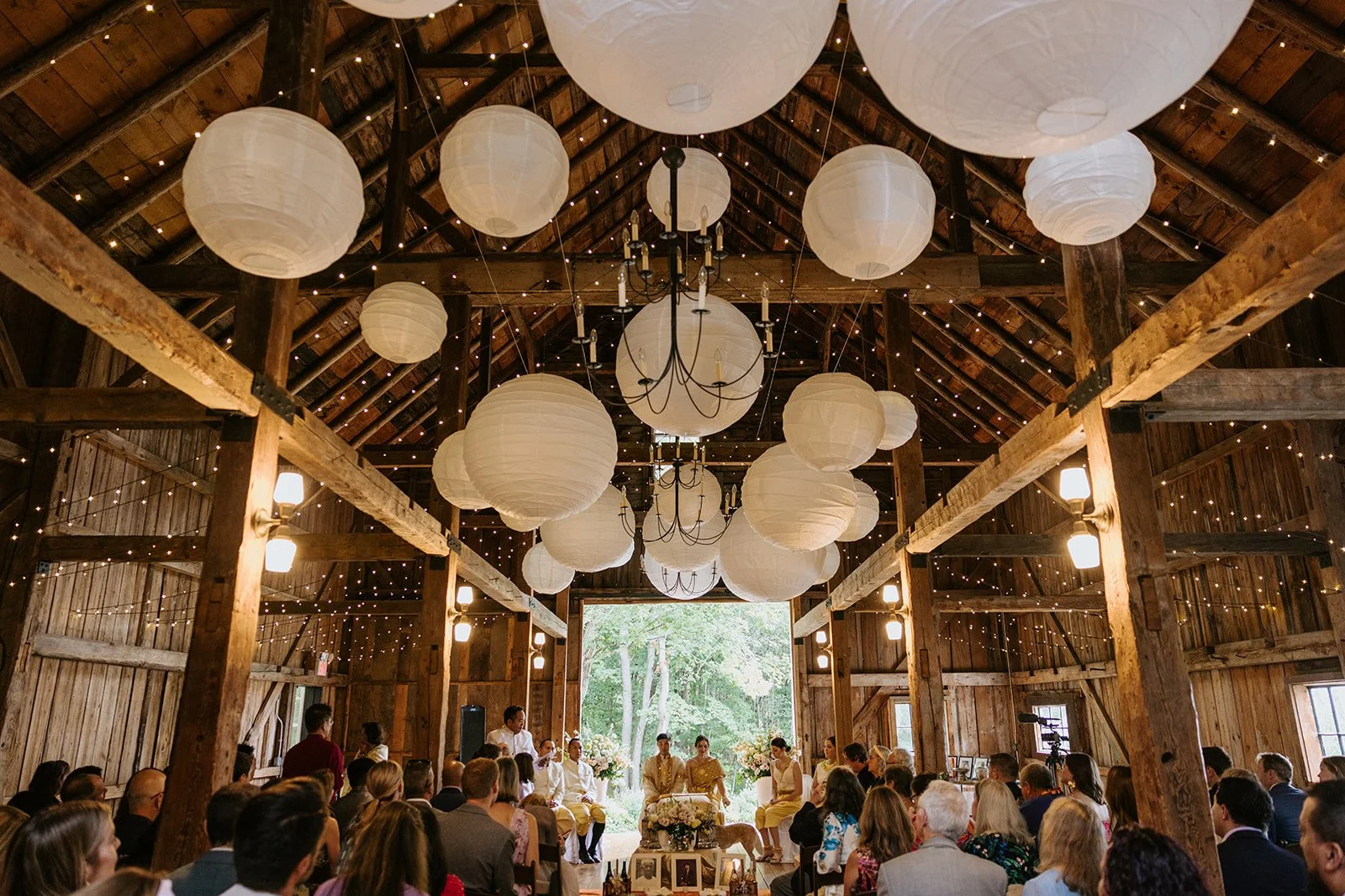 A rustic barn wedding ceremony with hanging white paper lanterns, string lights, and chandeliers, featuring seated guests and the wedding party at the altar.