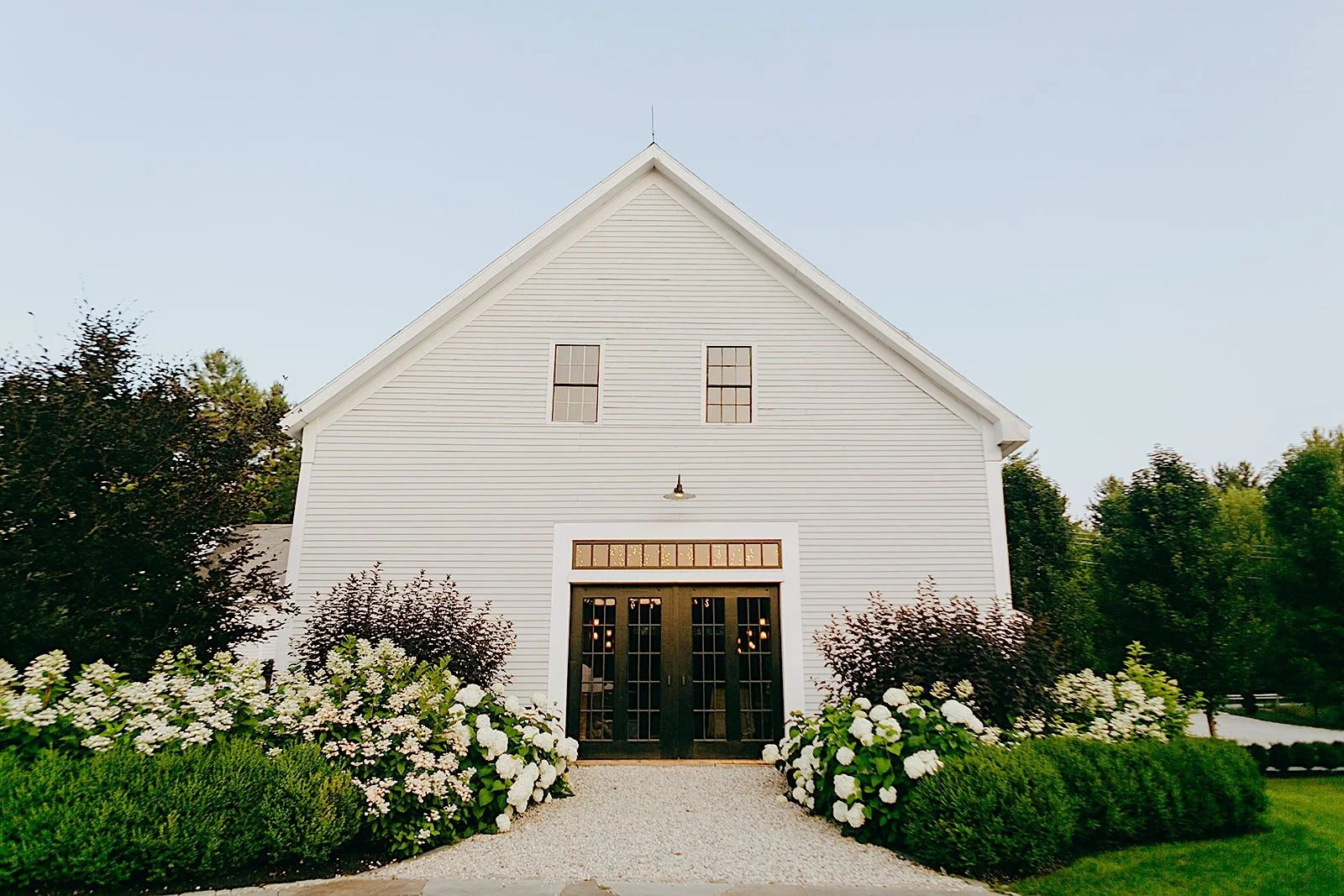 Front view of a white barn-style building with black door, surrounded by white flowers and greenery, under a clear blue sky.