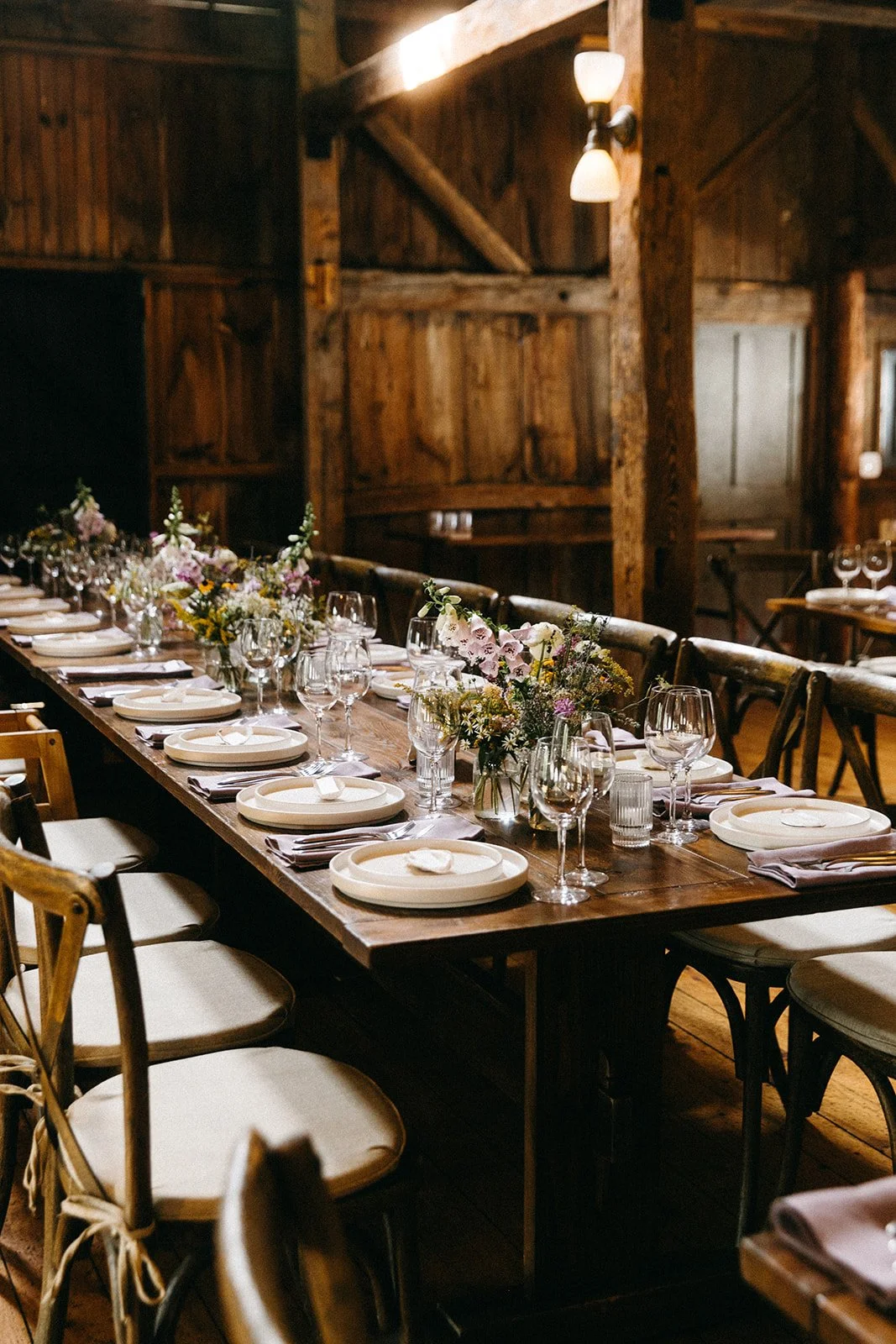 Long wooden dining table set for a formal event with white plates, wine glasses, and floral centerpieces, in a rustic wooden room with warm lighting.
