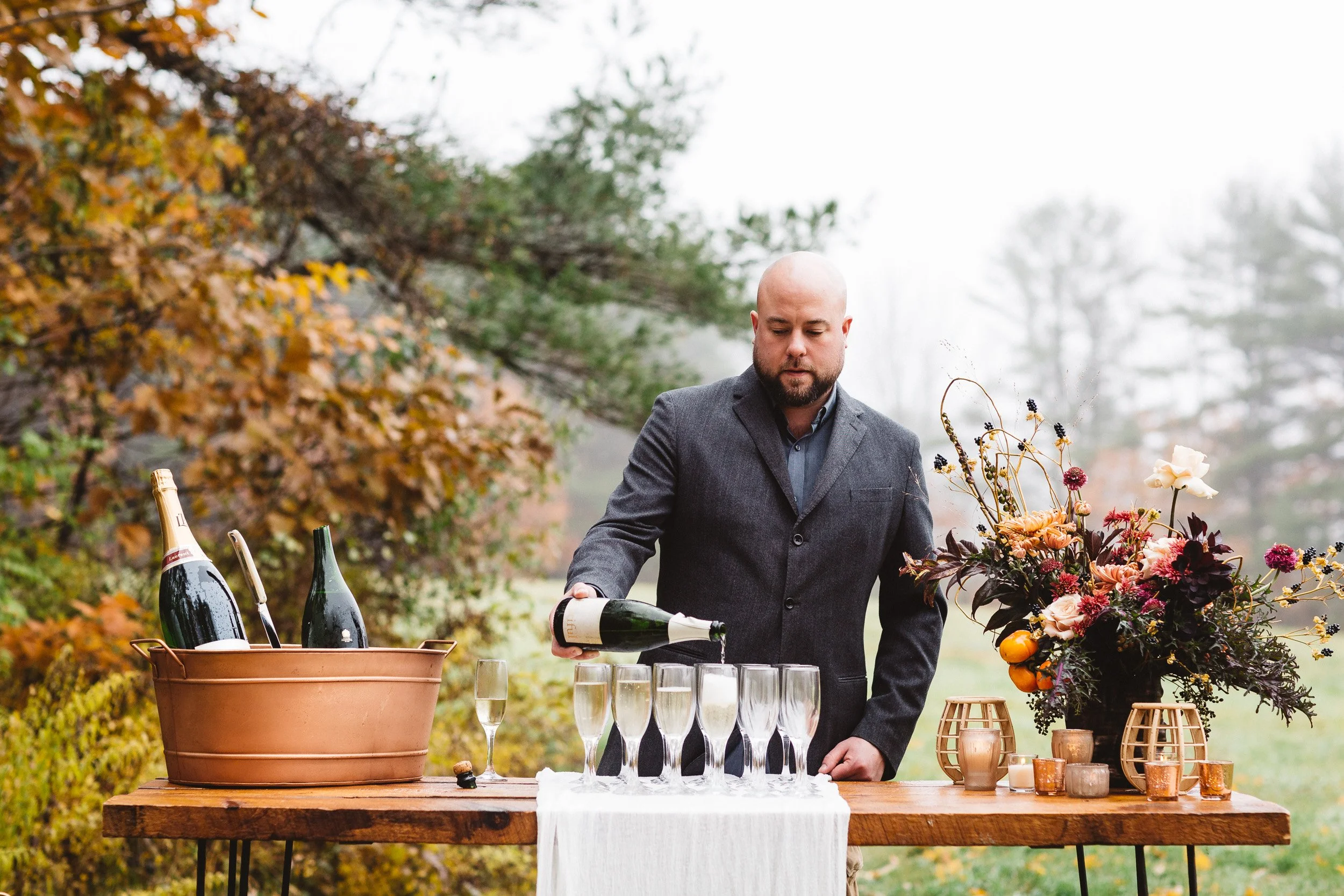 Man in gray suit pouring champagne into glasses on an outdoor table with floral arrangement and decorations, during fall.