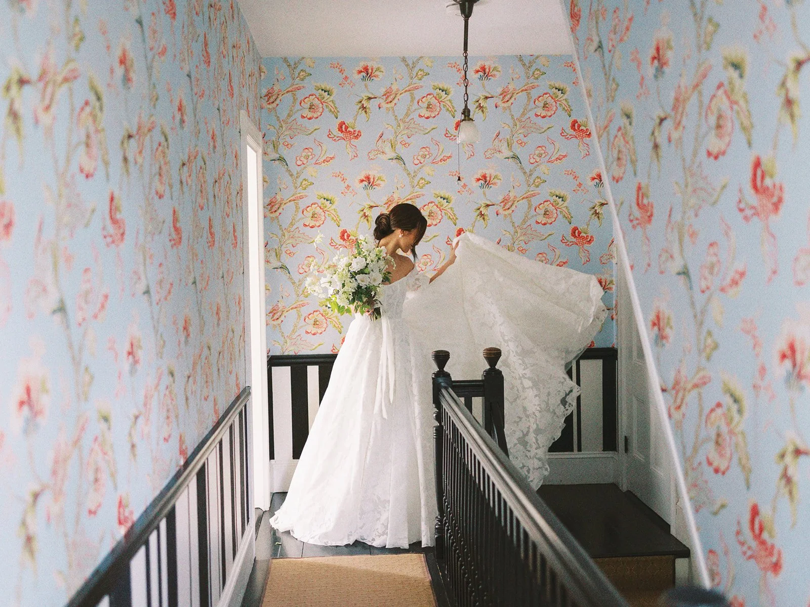 Brunette woman in wedding dress holding a bouquet of white flowers, walking upstairs in a narrow hallway with floral wallpaper.