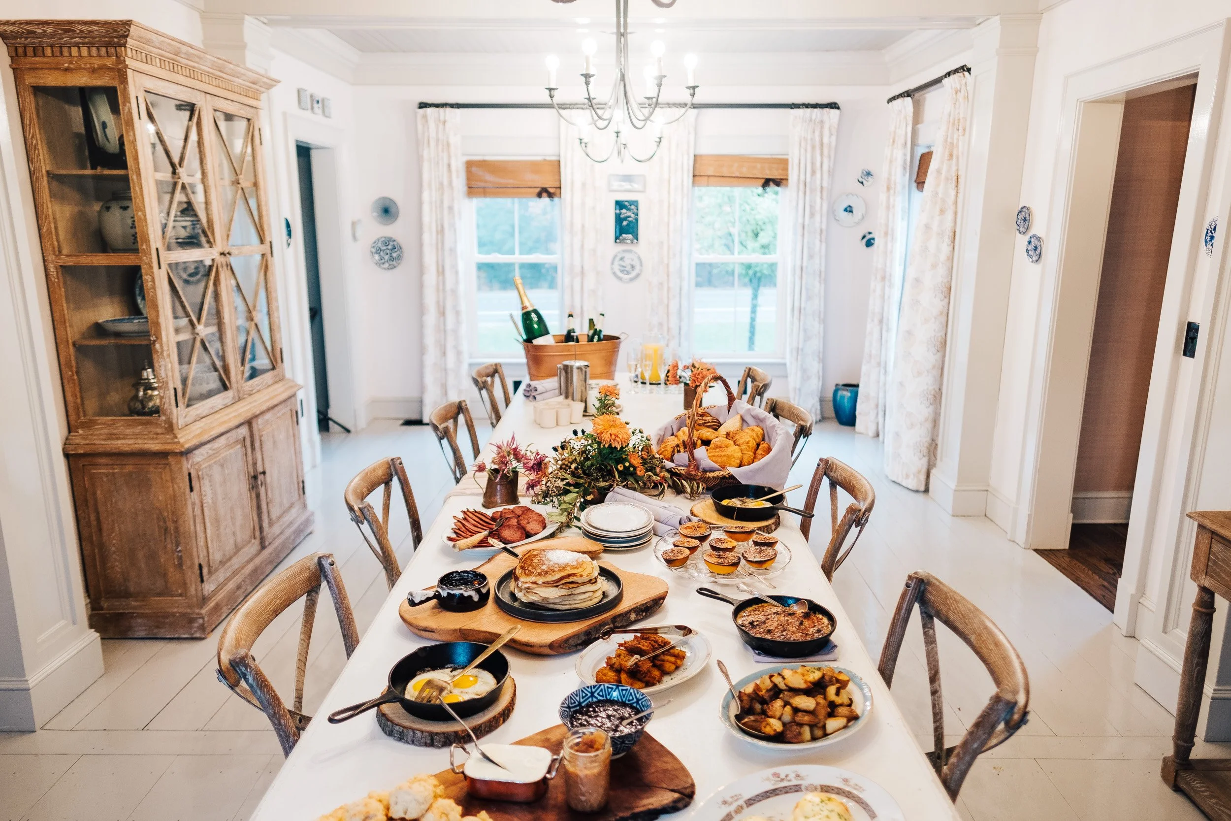 A dining room with a long table set for breakfast, featuring various dishes, breads, and drinks, with windows and curtains in the background and a wooden cabinet on the side.