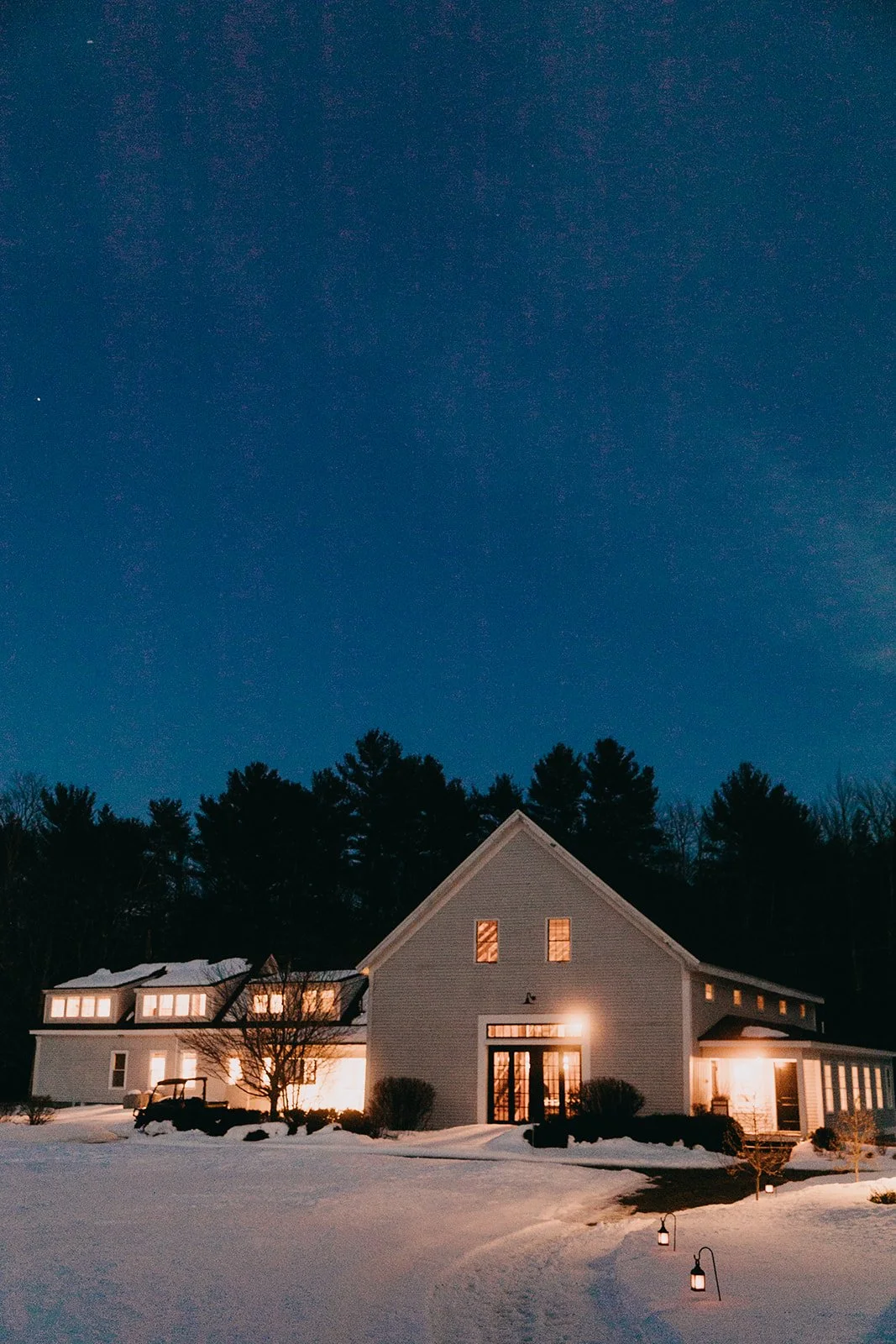 Snow-covered house at night with warm lights, surrounded by trees and outdoor lanterns along a snow path.