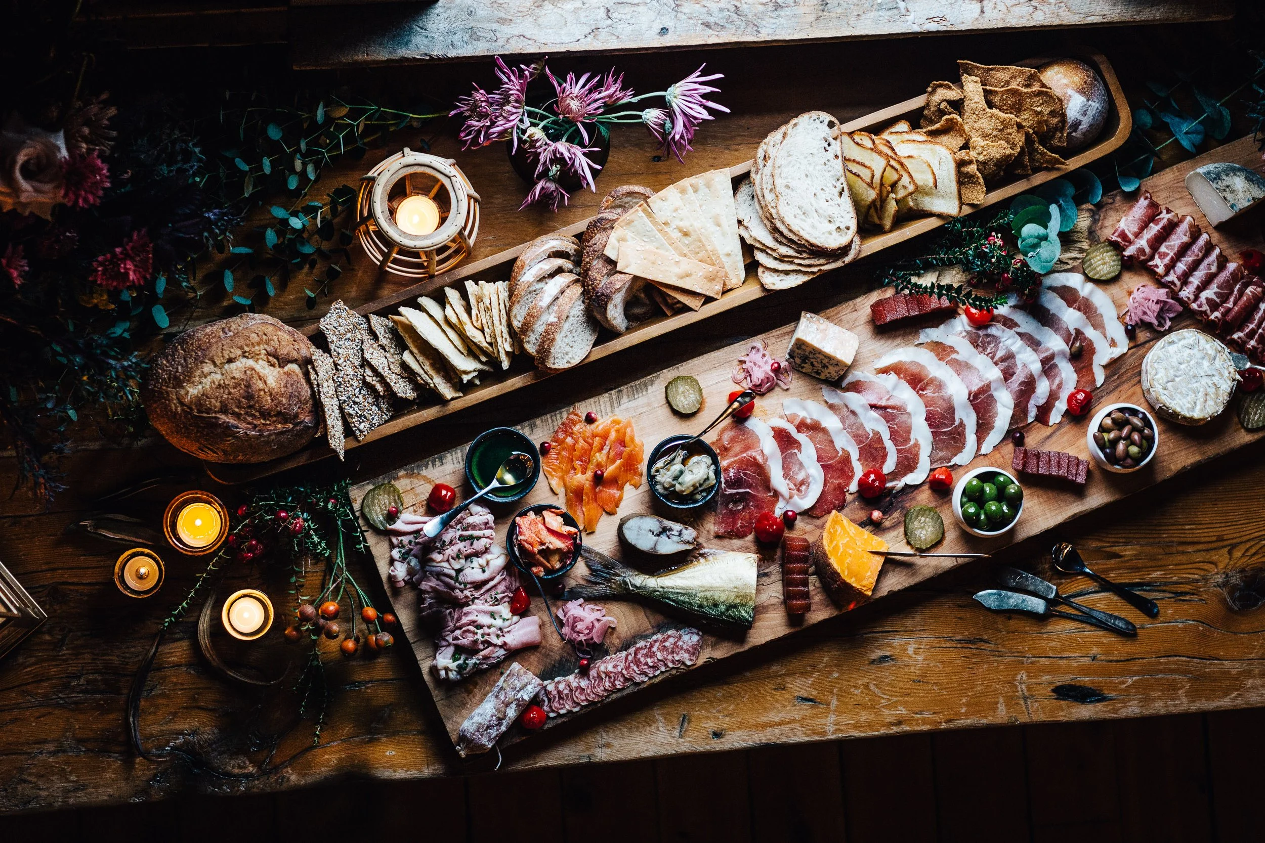 A festive charcuterie board with cheese, meats, bread, crackers, pickles, and accompaniments on a wooden table, decorated with candles, flowers, and greenery.