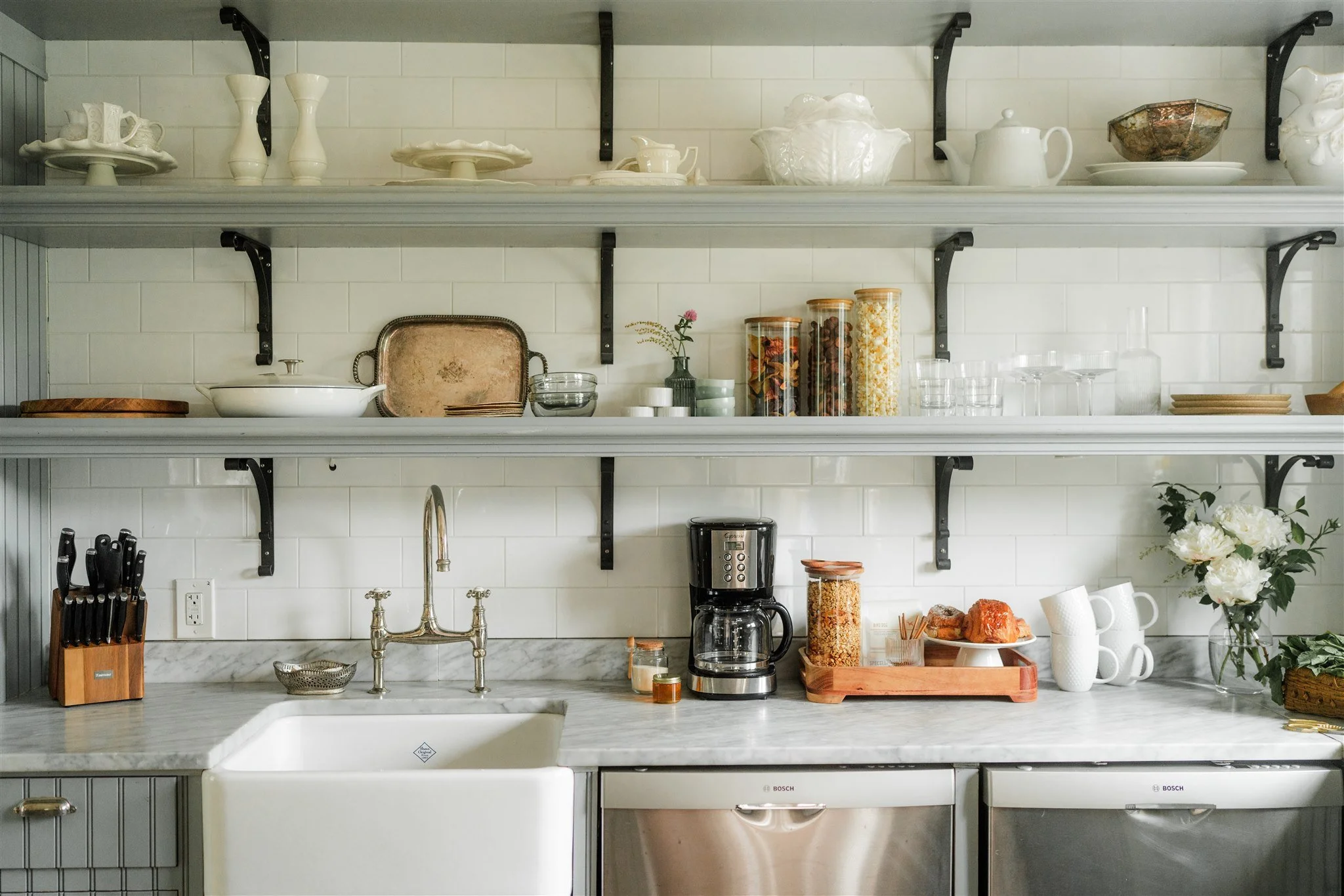 Kitchen with white subway tile backsplash, open shelves with dishes, jars of food, coffee maker, knives, flowers, and a farmhouse sink.