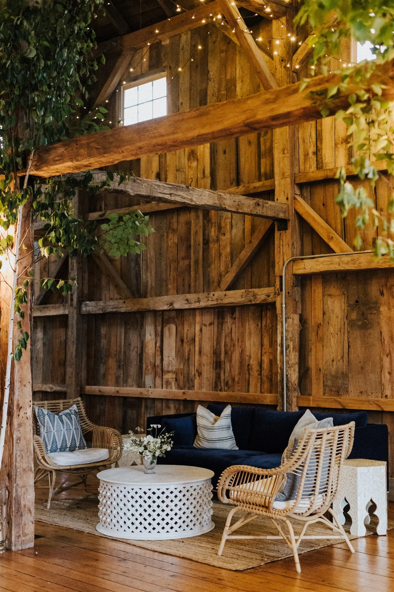 Cozy rustic indoor space with wooden walls and ceiling, decorated with string lights, featuring a black sofa, wicker chairs, a white coffee table with a flower arrangement, and cushions.